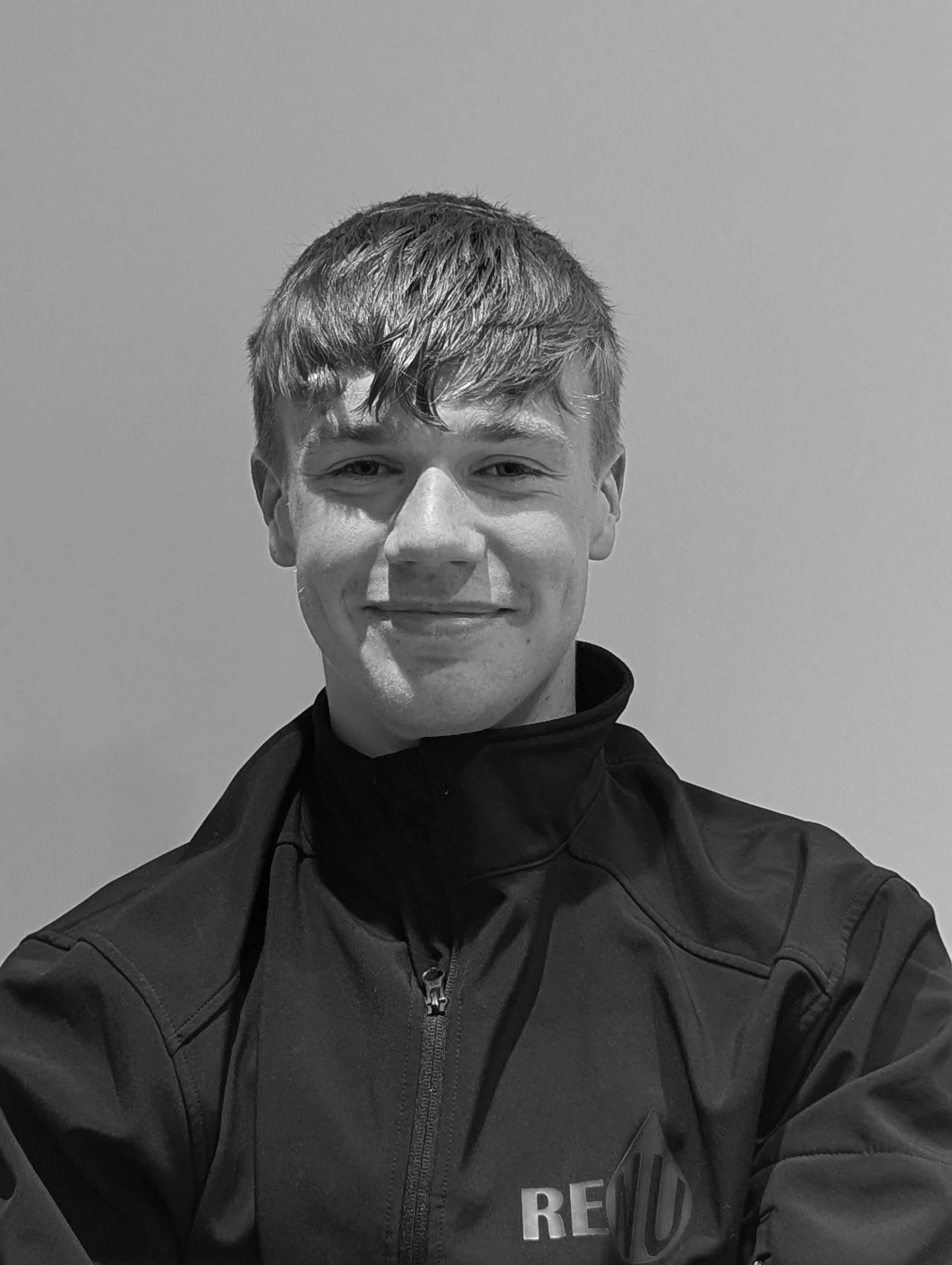 A young man with short, wavy hair smiling, wearing a zip-up jacket, against a plain wall.