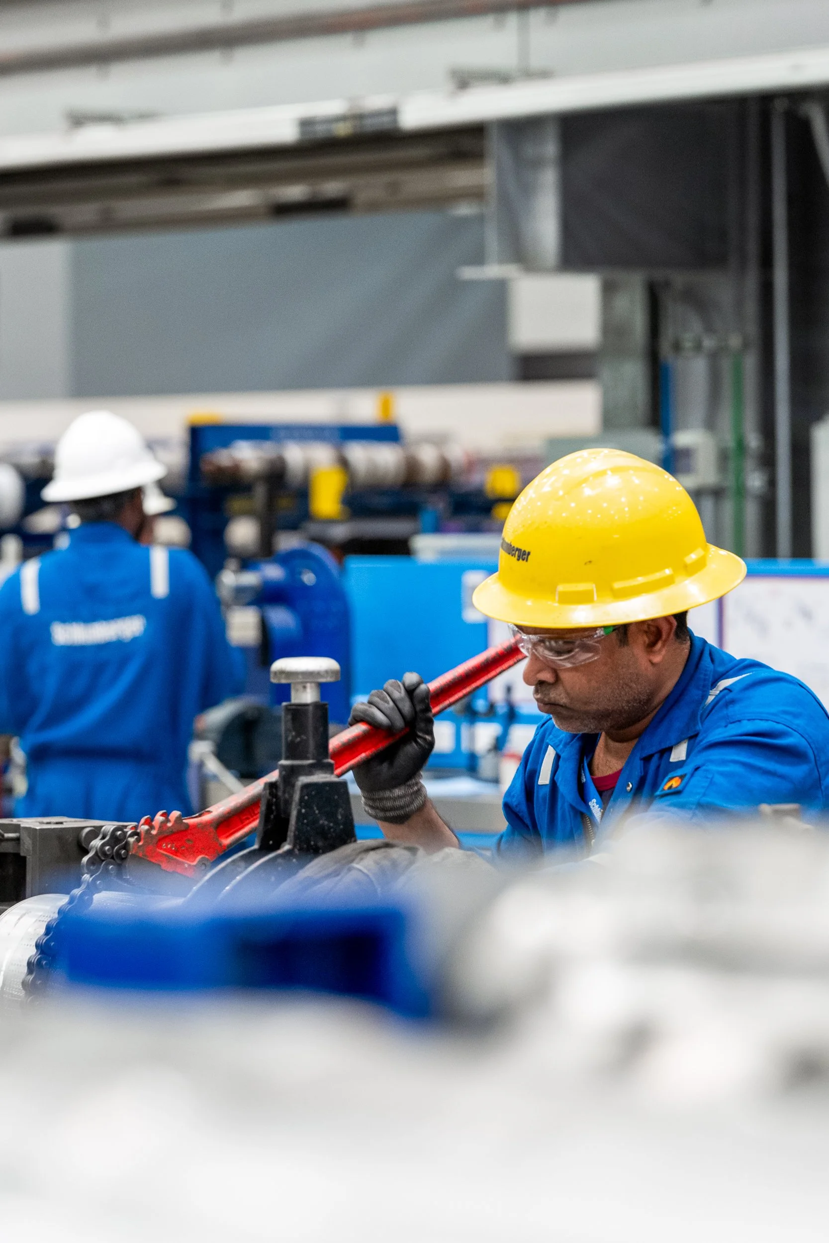 An industrial worker wearing a yellow safety helmet, safety goggles, and gloves operating a machine with a red handle in a factory setting, with other workers in the background.