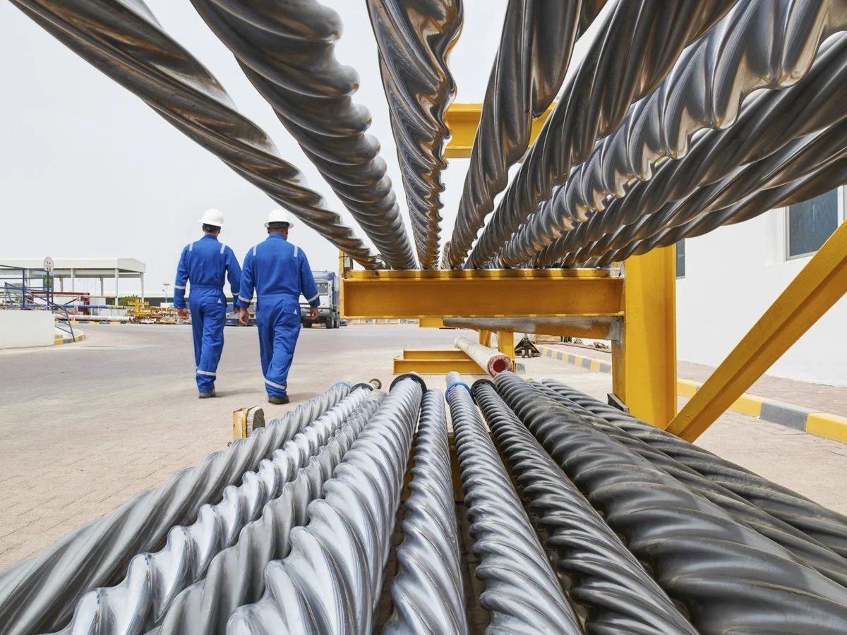 Two workers in blue coveralls and white helmets walking past large industrial cables or pipes on a yellow storage rack outside a facility.