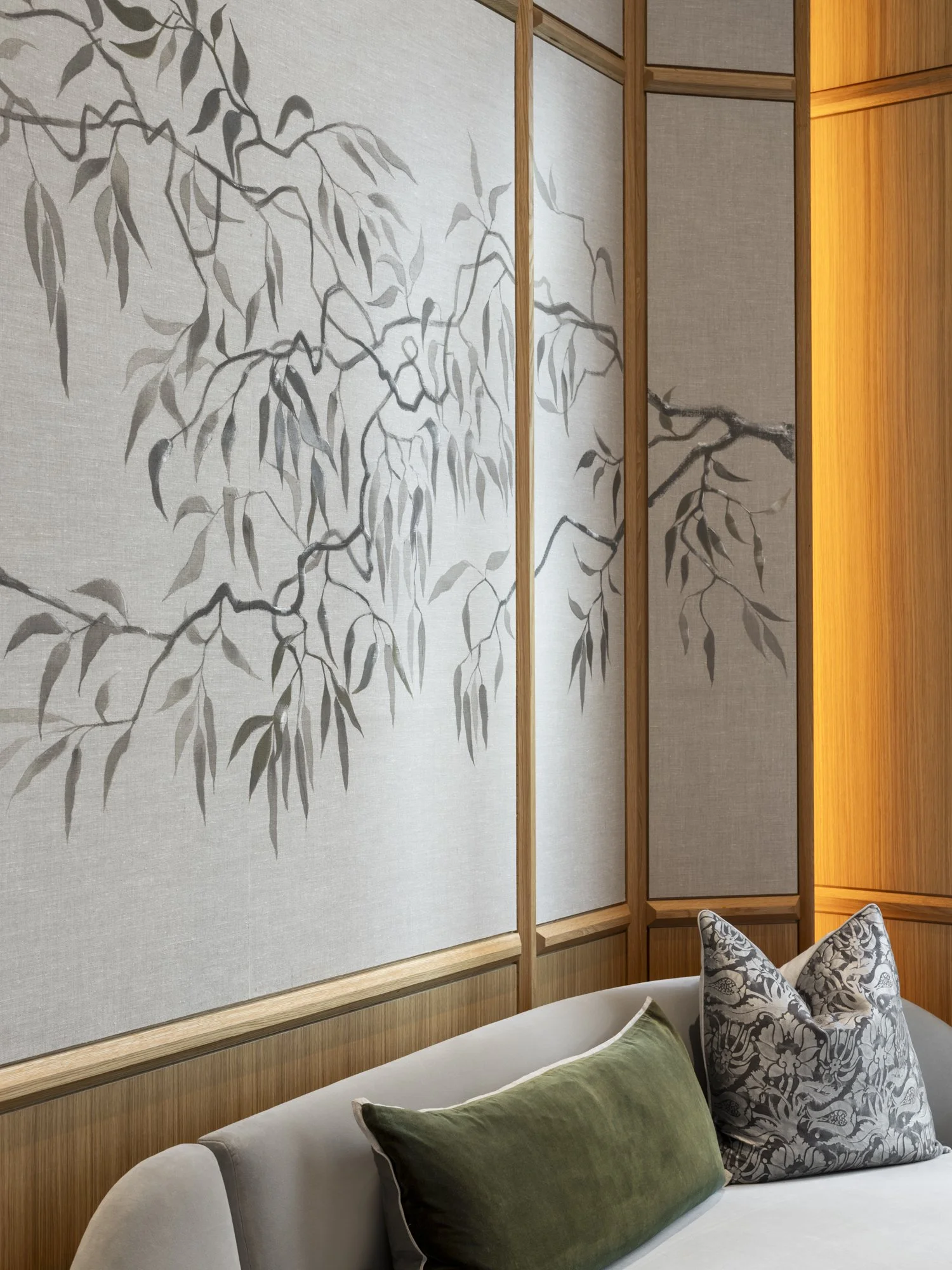 A wall with a large, artistic panel featuring a painted or printed branch with leaves, decorated with wooden trim. A white sofa with green and patterned pillows is in front of the wall.