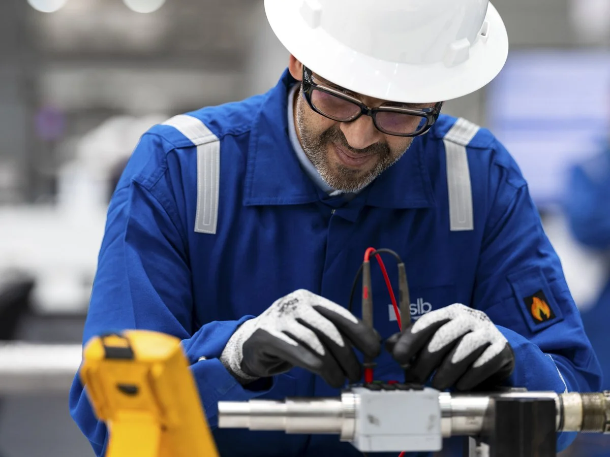 A worker wearing a hard hat, safety glasses, and gloves is working on an industrial machine or equipment, attaching or fixing wires in a factory or workshop.