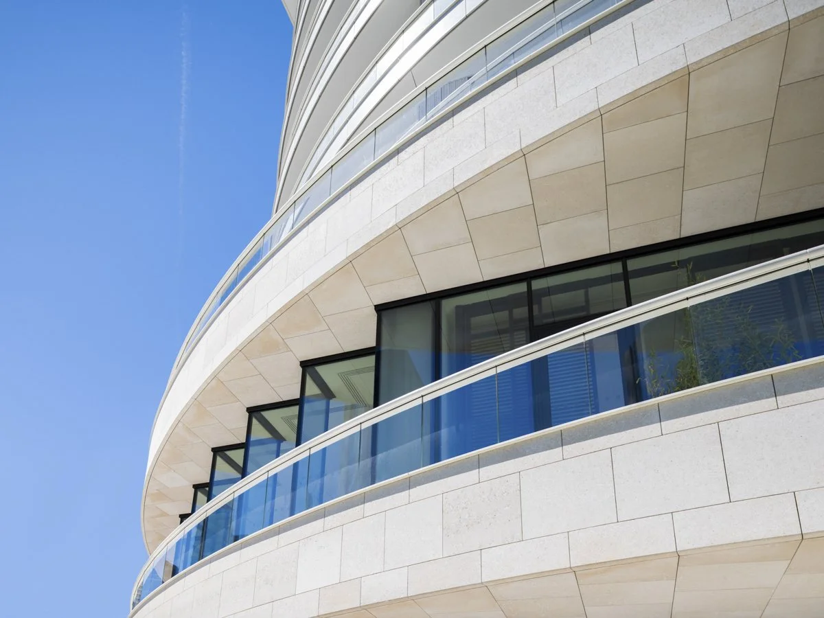Close-up of a modern building with curved white exterior walls, glass windows, and blue railings, against a clear blue sky.
