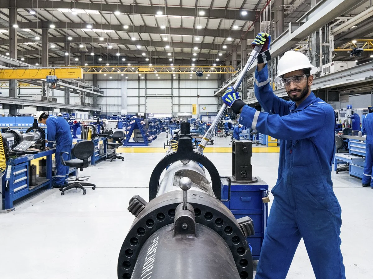 A worker in blue coveralls and a white safety helmet using a tool on a large industrial pipe inside a spacious manufacturing facility with multiple workers and equipment.
