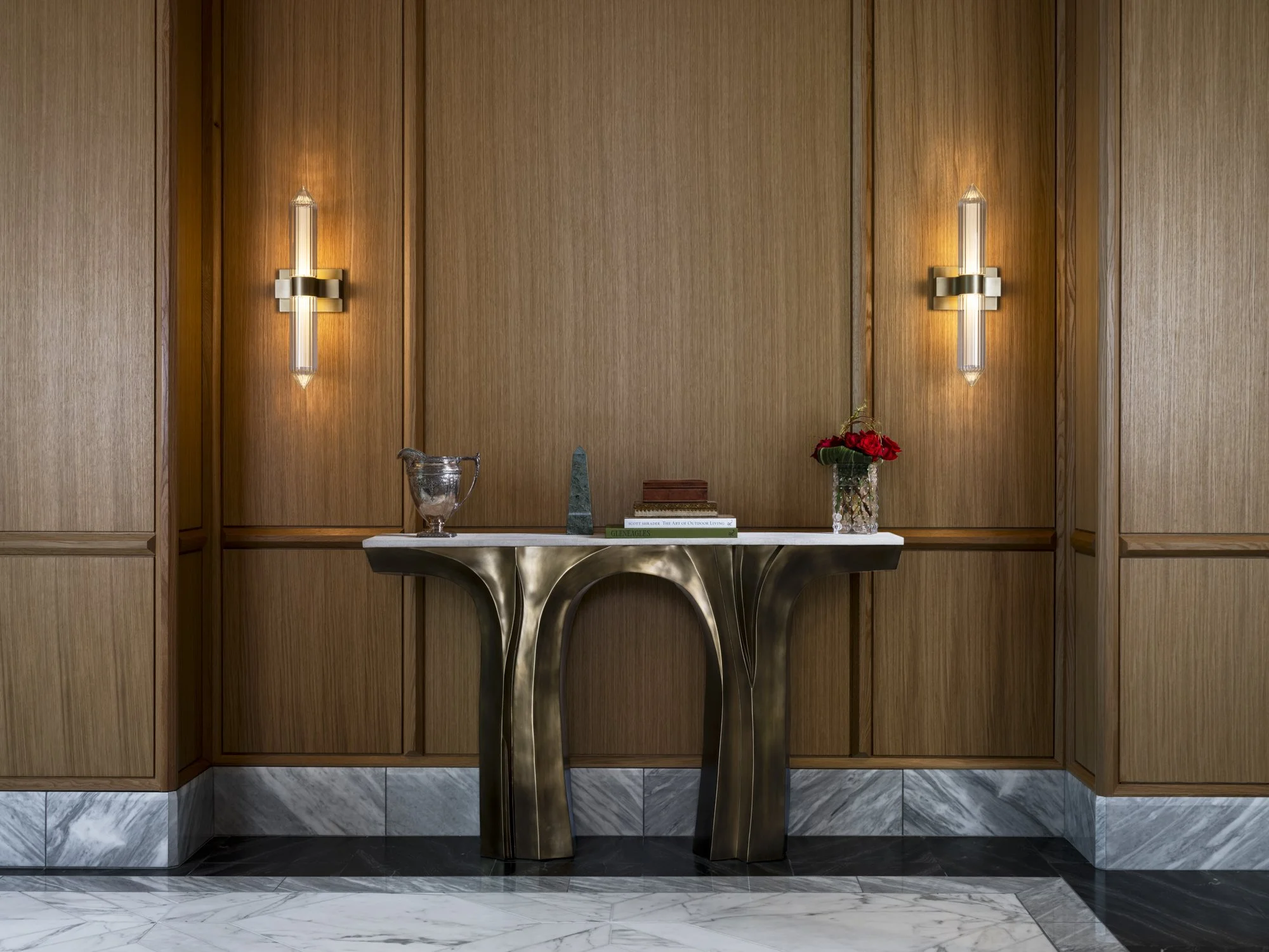 Wood-paneled wall with two modern wall sconces, a marble-topped side table with decorative items including a silver pitcher, a green sculpture, a stack of books, and a vase of red roses.