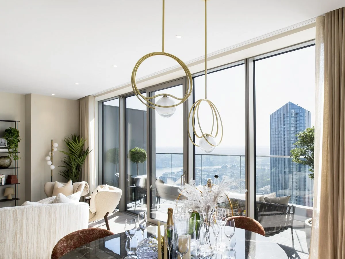 Bright modern living room with large glass windows, outdoor balcony, and city view, decorated with plants and elegant furniture.