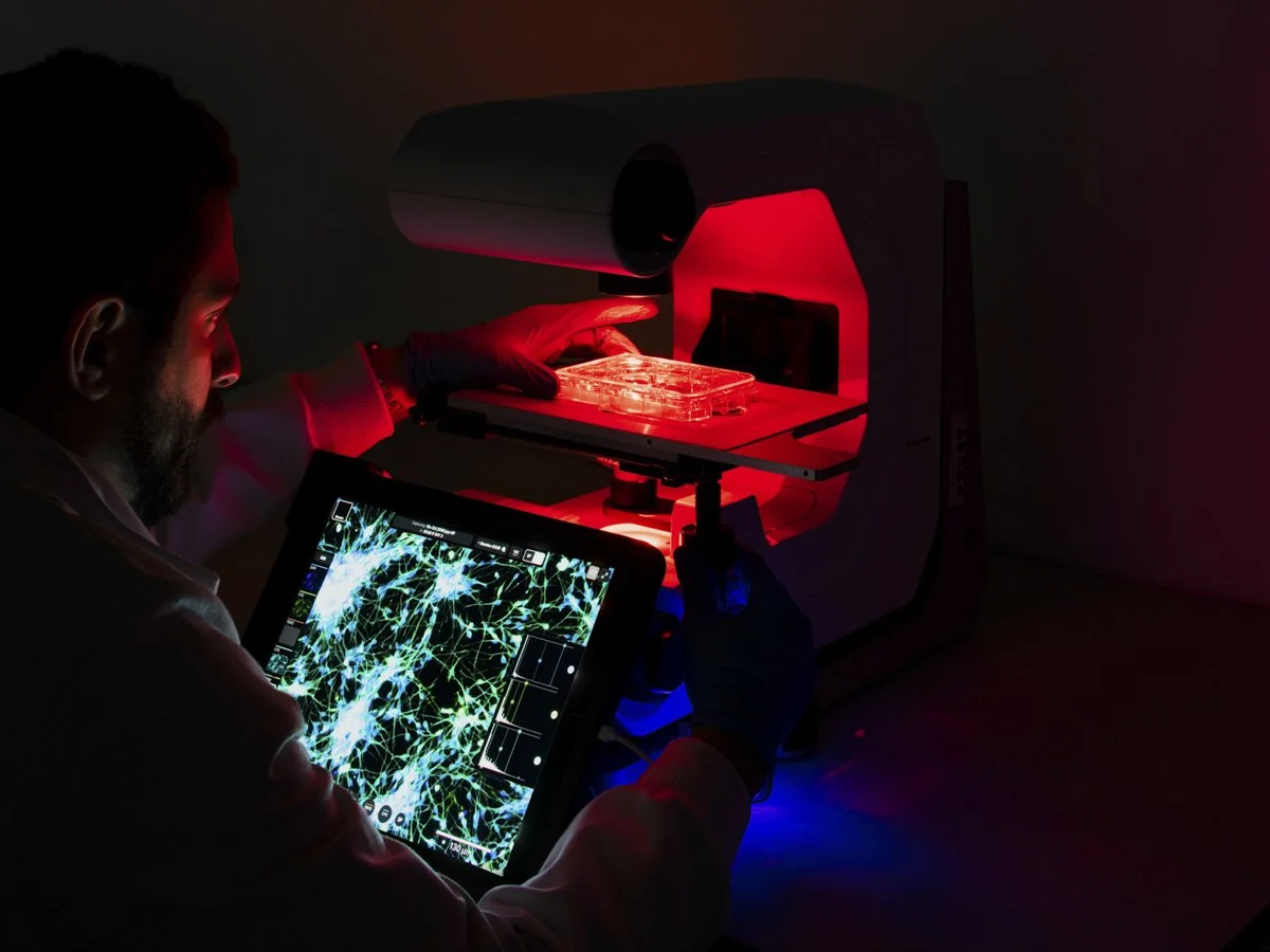 Scientist using a microscope with a red light to analyze a sample, with a digital screen displaying neural network imagery.