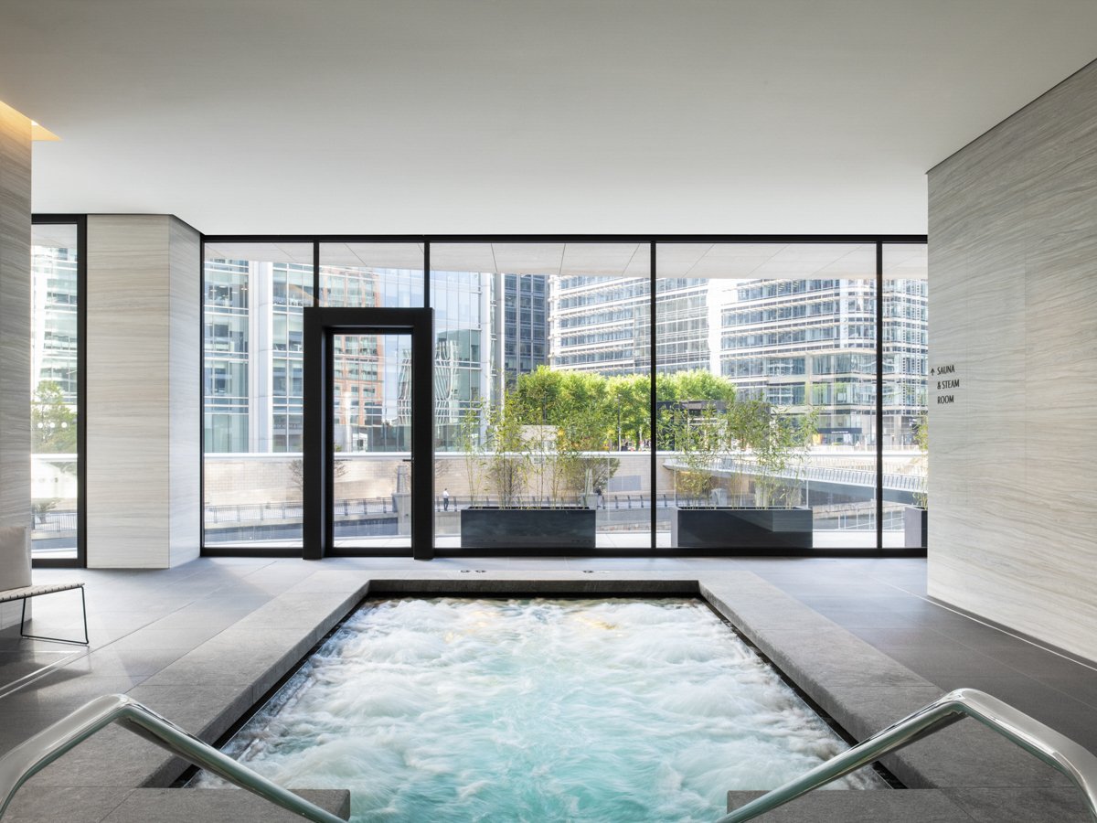 Indoor spa with a bubbling hot tub in front of large floor-to-ceiling windows showing a cityscape with tall modern buildings and some green trees outside.