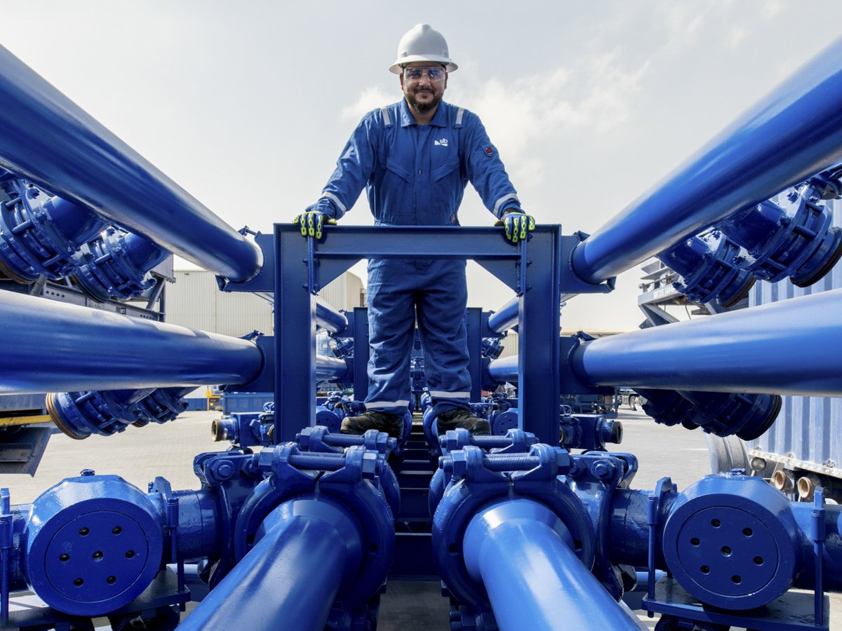 A man wearing safety gear, including a hard hat, safety glasses, and gloves, standing on a blue industrial platform surrounded by large blue pipes in an industrial setting.