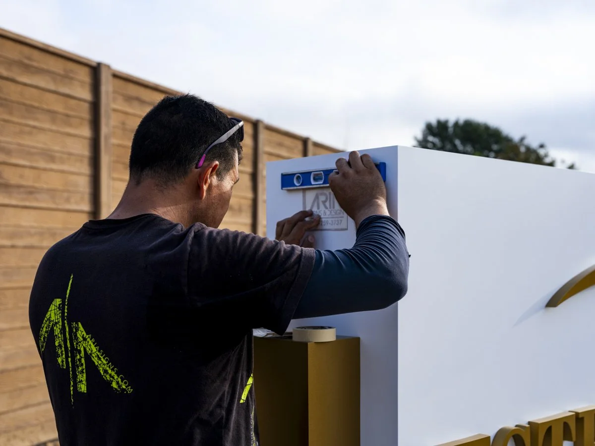 A man working on a white signboard outdoors with a wooden fence in the background. He is attaching a sticker or label to the board, wearing a black shirt and sunglasses on his head.