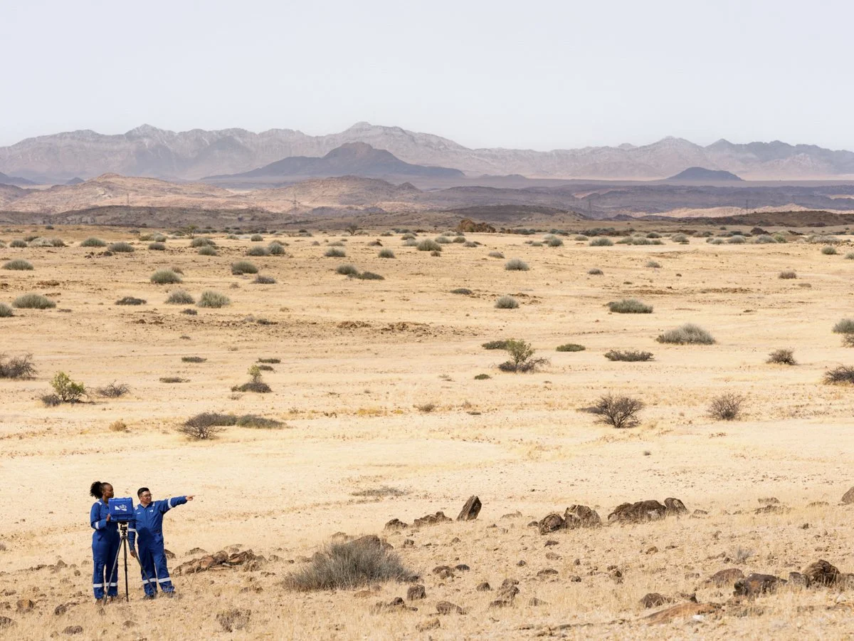 Two scientists in blue uniforms working in a vast desert with rocks and sparse vegetation, mountains in the background.