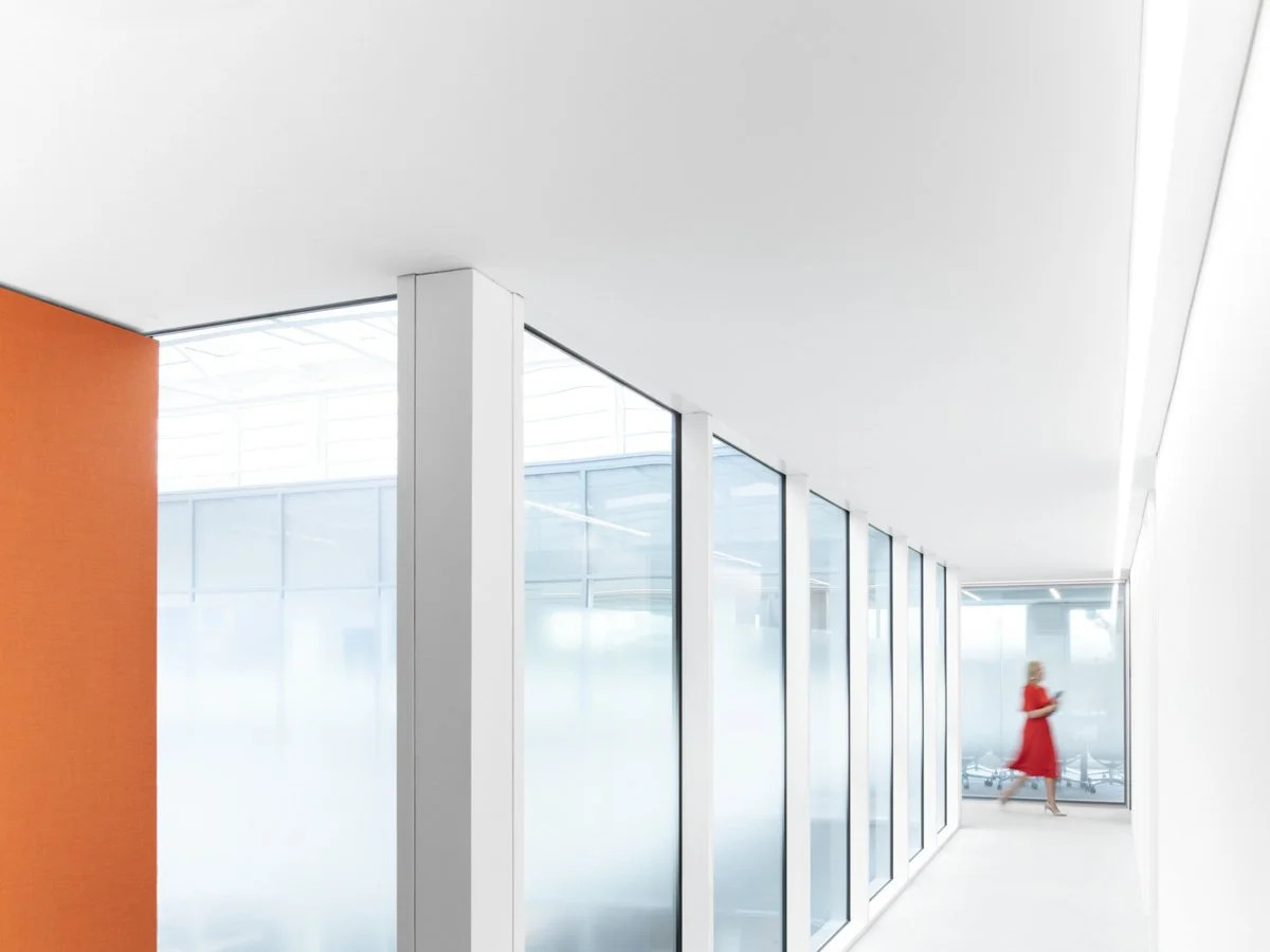 Modern, minimalistic indoor hallway with white walls and ceiling, large glass windows, and an orange accent wall on the left. A woman in a red dress is walking in the background.