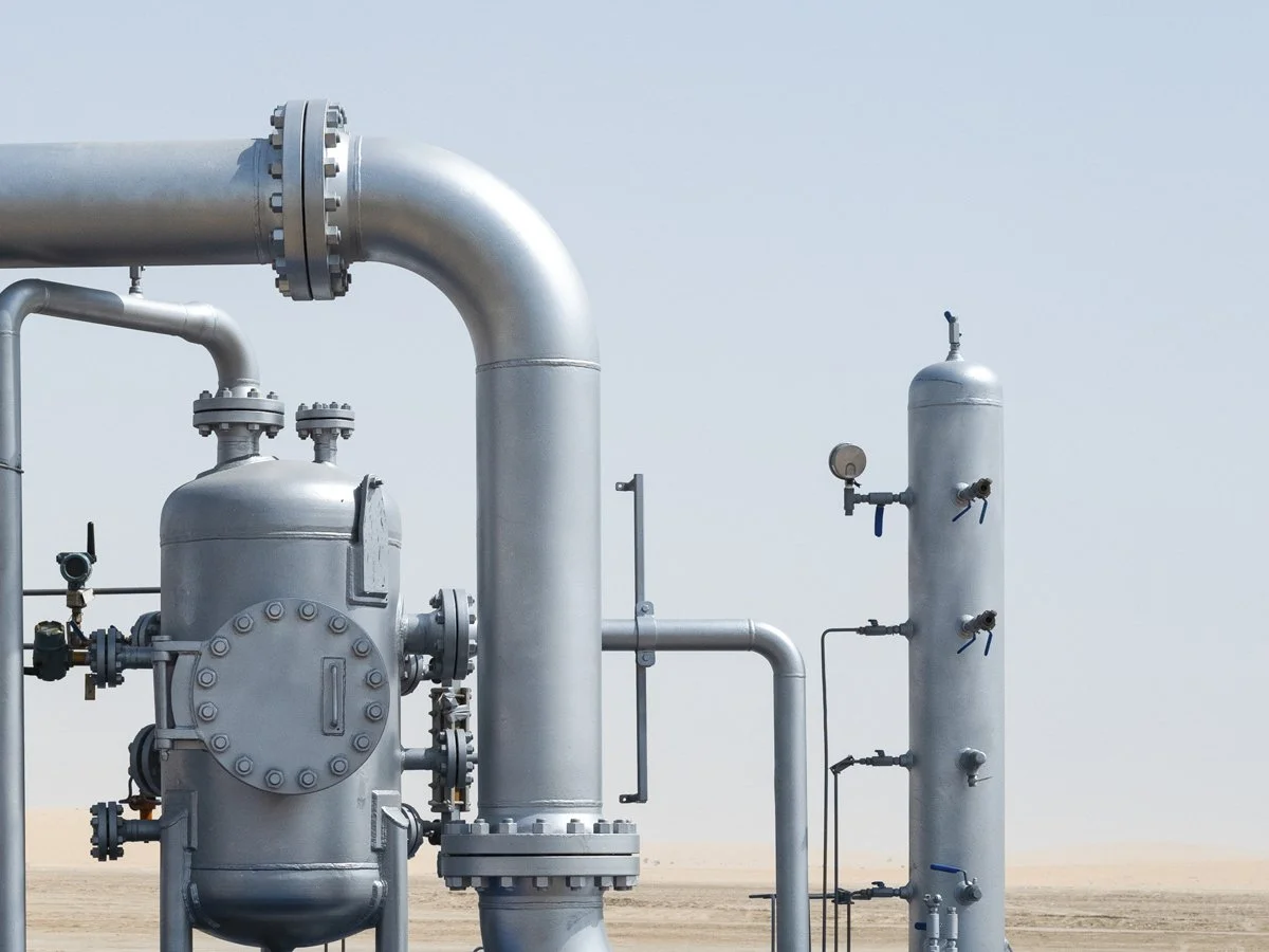 Industrial pipes, valves, and pressure gauges in a desert setting under a blue sky.