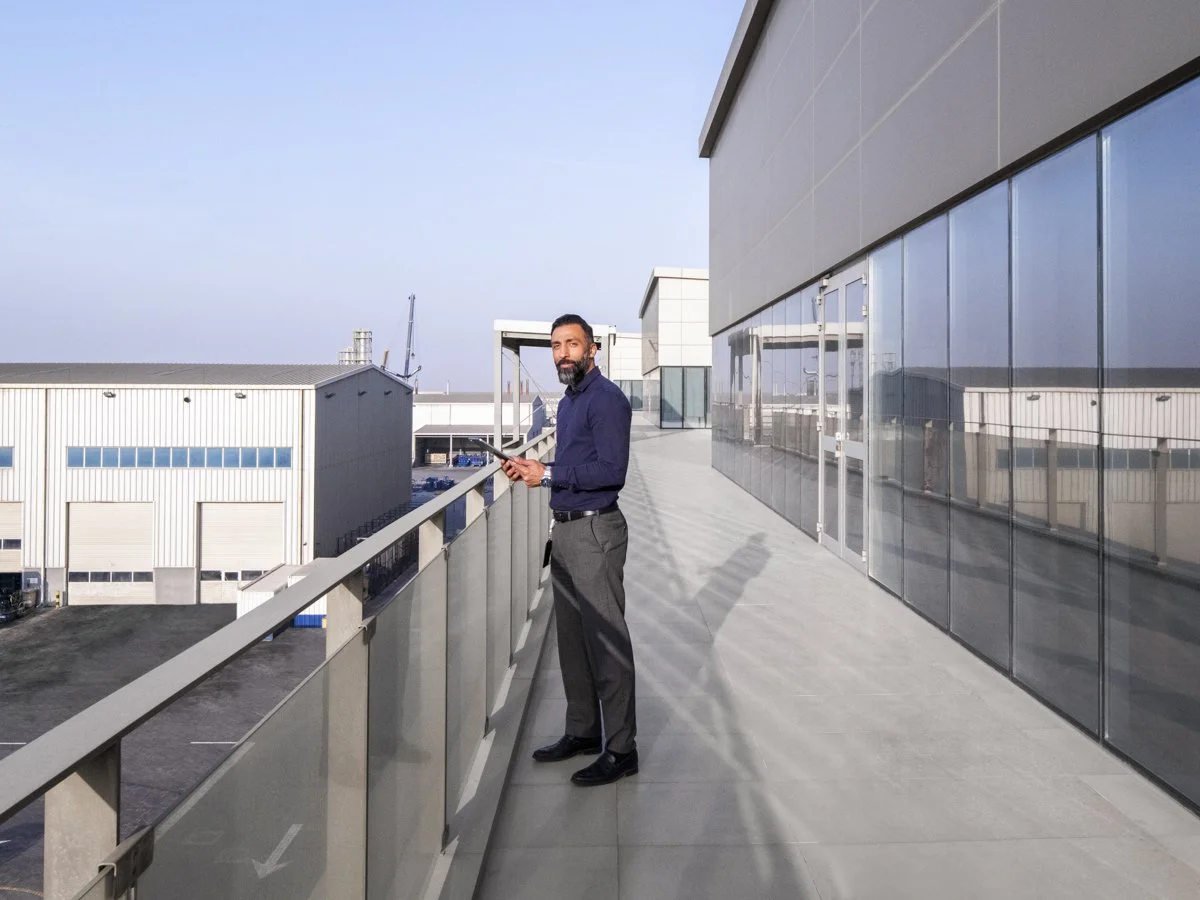 A man with a beard wearing a dark blue shirt and gray pants holding a tablet on a rooftop balcony of a modern building with reflective glass windows, with industrial buildings and a clear sky in the background.