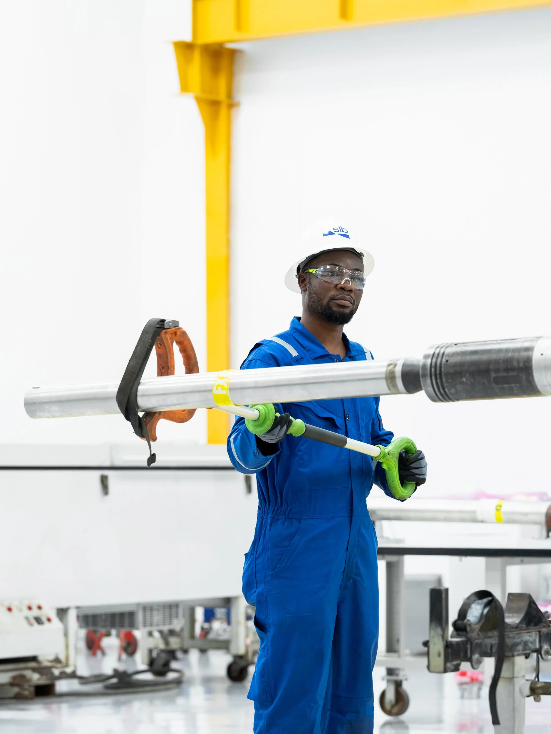 A worker in blue overalls, safety glasses, and a white hard hat holds a long metal pipe with a green clamp in a manufacturing or industrial setting.