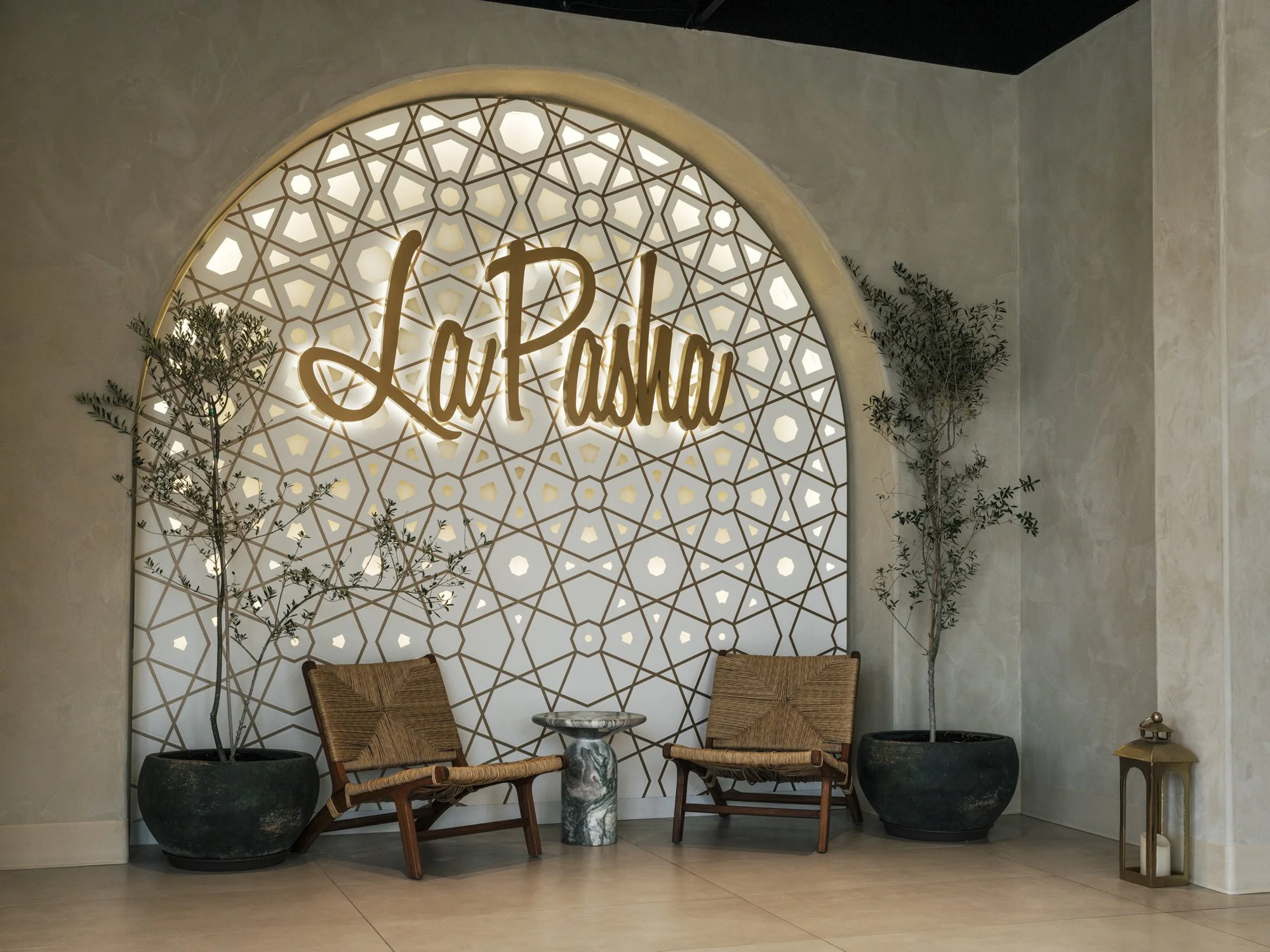 Decorative wall with a glowing sign that says "La Pasha," framed by intricate geometric patterns. Two potted trees and two wooden chairs with woven seats and backs are arranged in front, with a small marble-topped side table between them. A lantern i