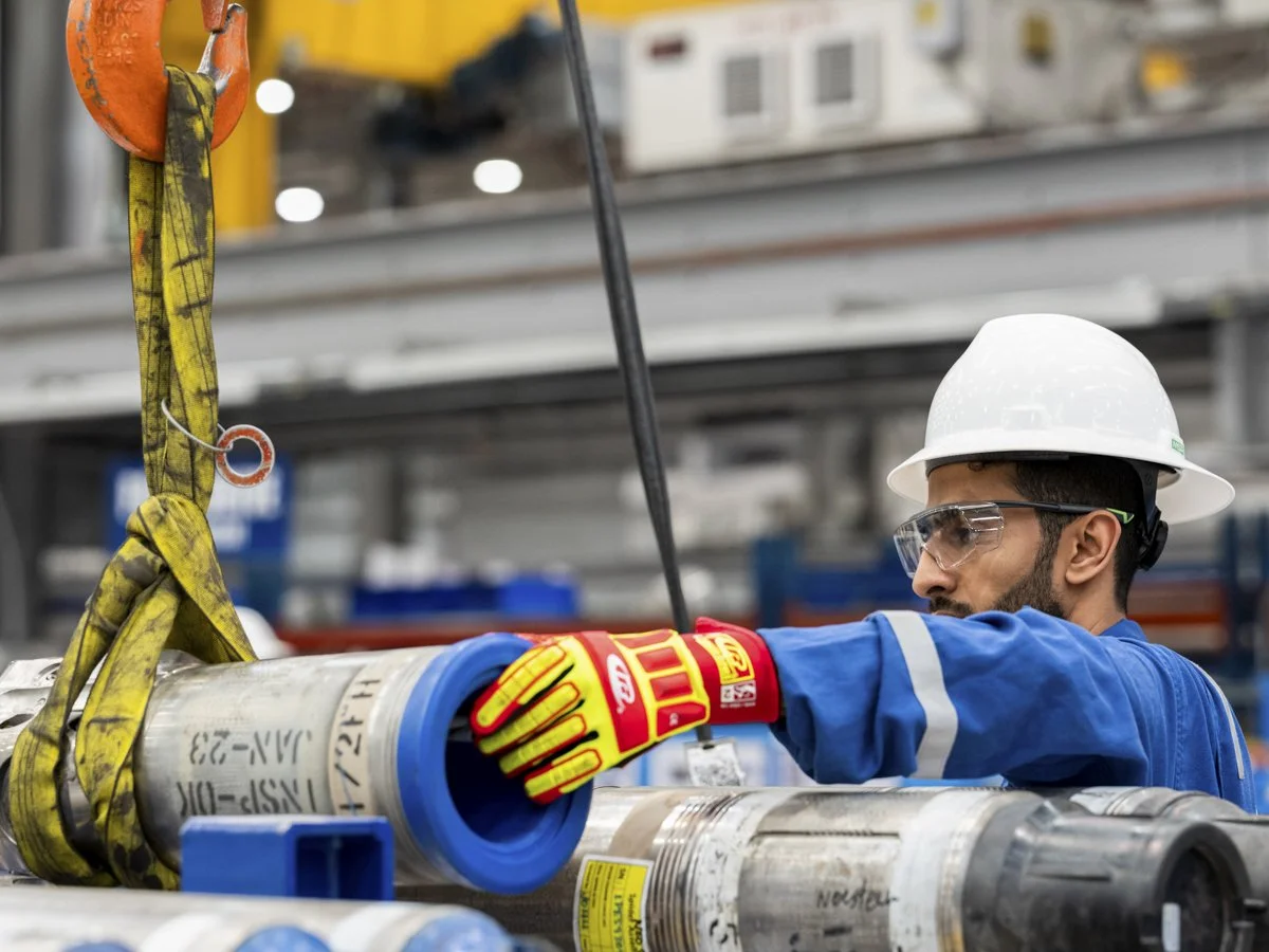 A construction worker wearing a white safety helmet, clear safety glasses, yellow work gloves, and a blue uniform working on industrial pipes with a yellow and black hoist strap in an industrial setting.