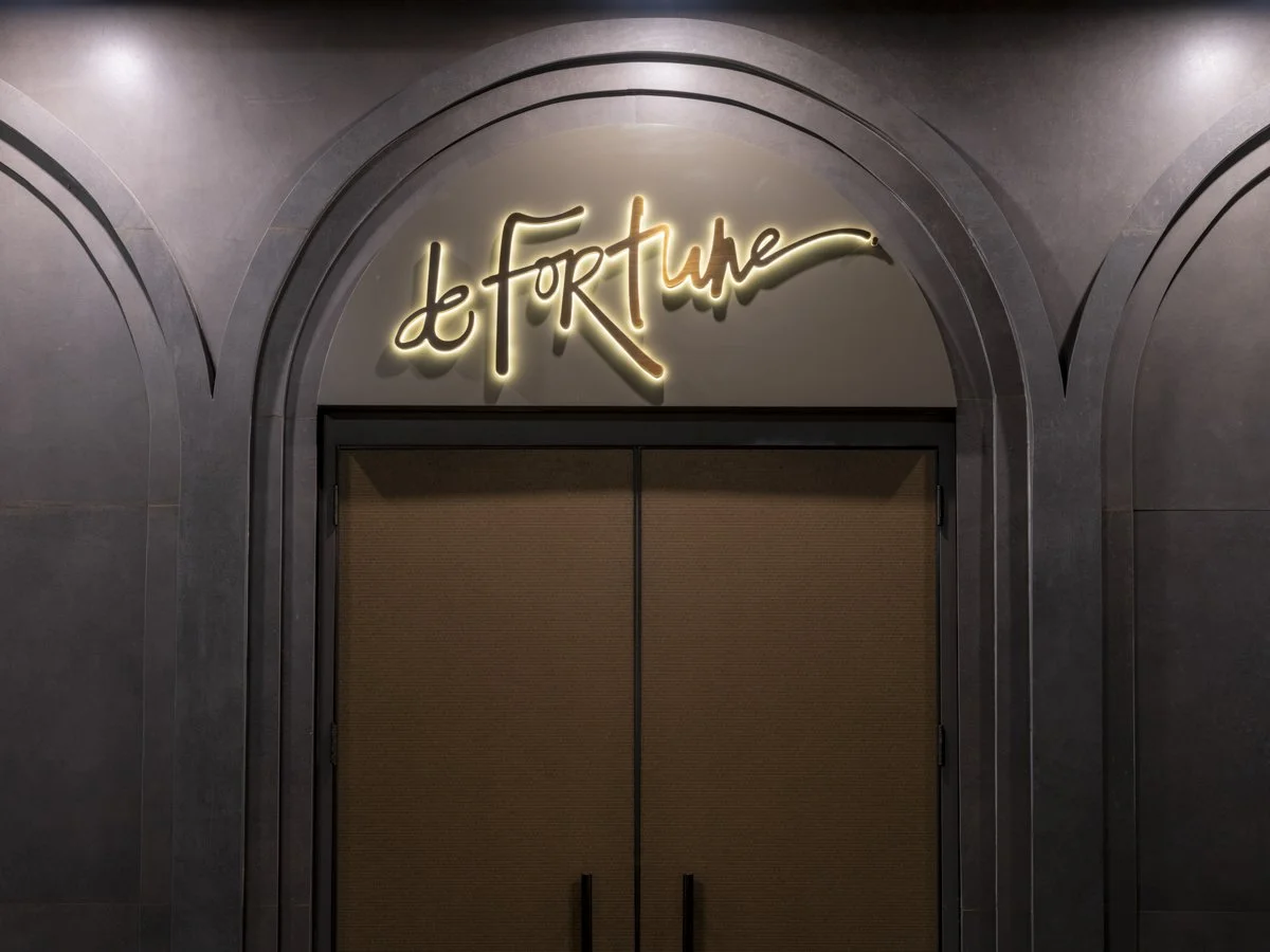 Neon sign above a doorway spelling 'Le Fortune' in cursive, with dark interior doors below, in an arched wall setting.