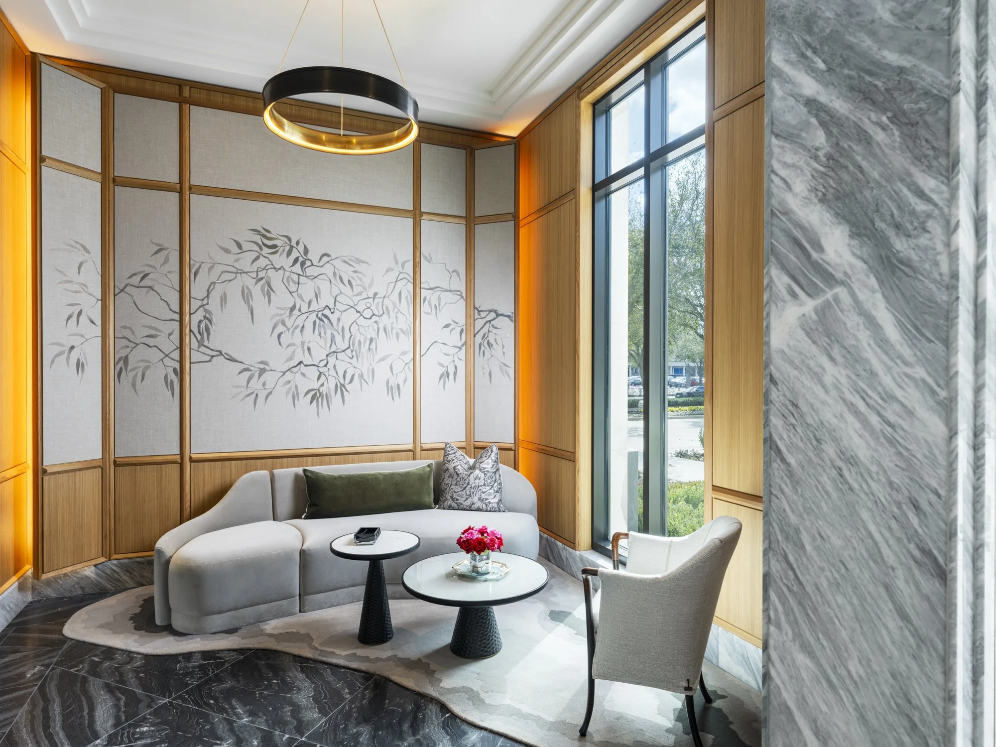 A modern, elegantly decorated lounge area with a white curved sofa, a small round black coffee table with a vase of pink flowers, a single white armchair, and a large window letting in natural light. The walls feature wooden paneling and a gray marbl