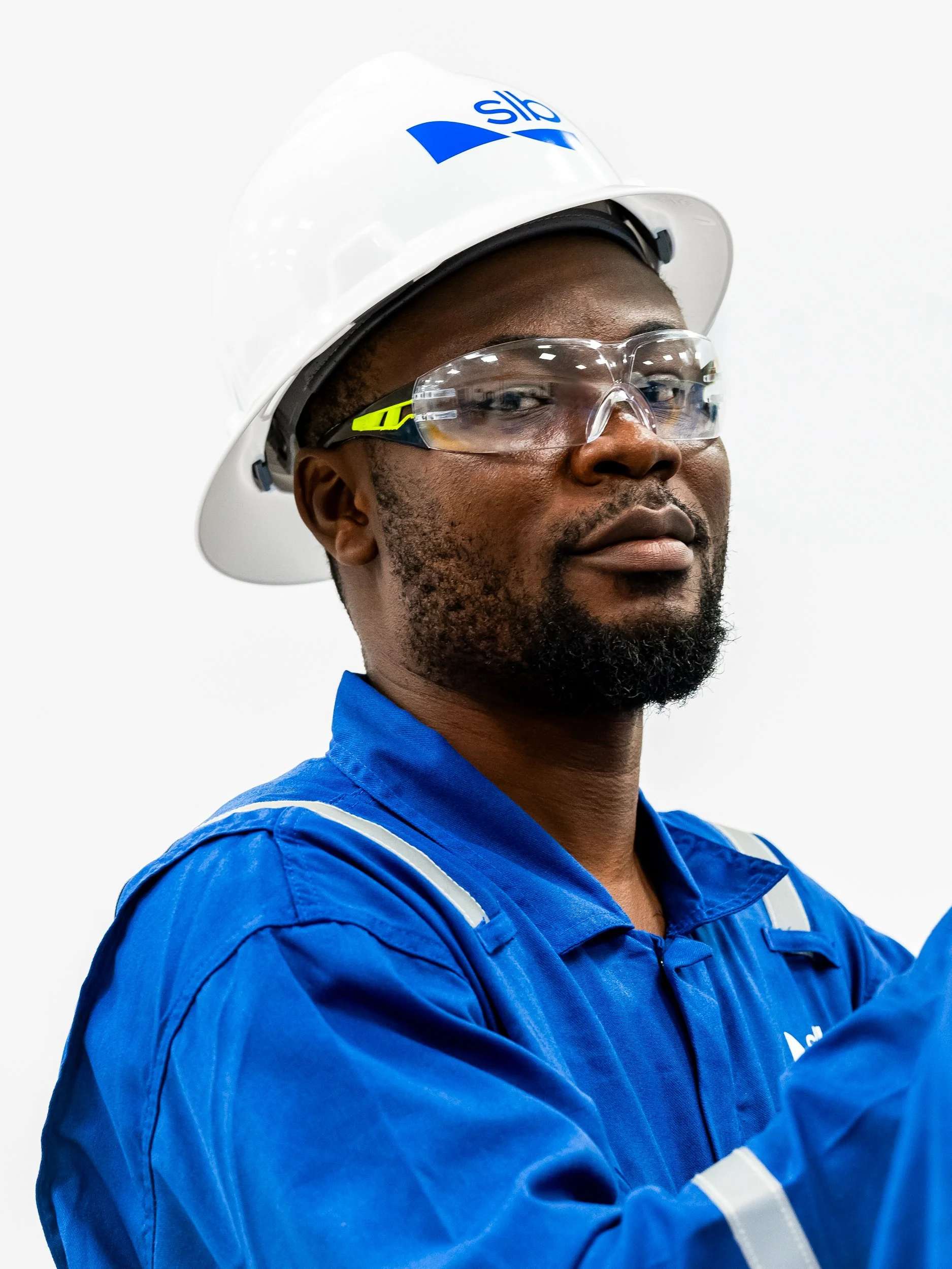 Close-up of an African American man wearing safety glasses, a white hard hat with a blue logo, and a blue work uniform, looking confidently to the side.