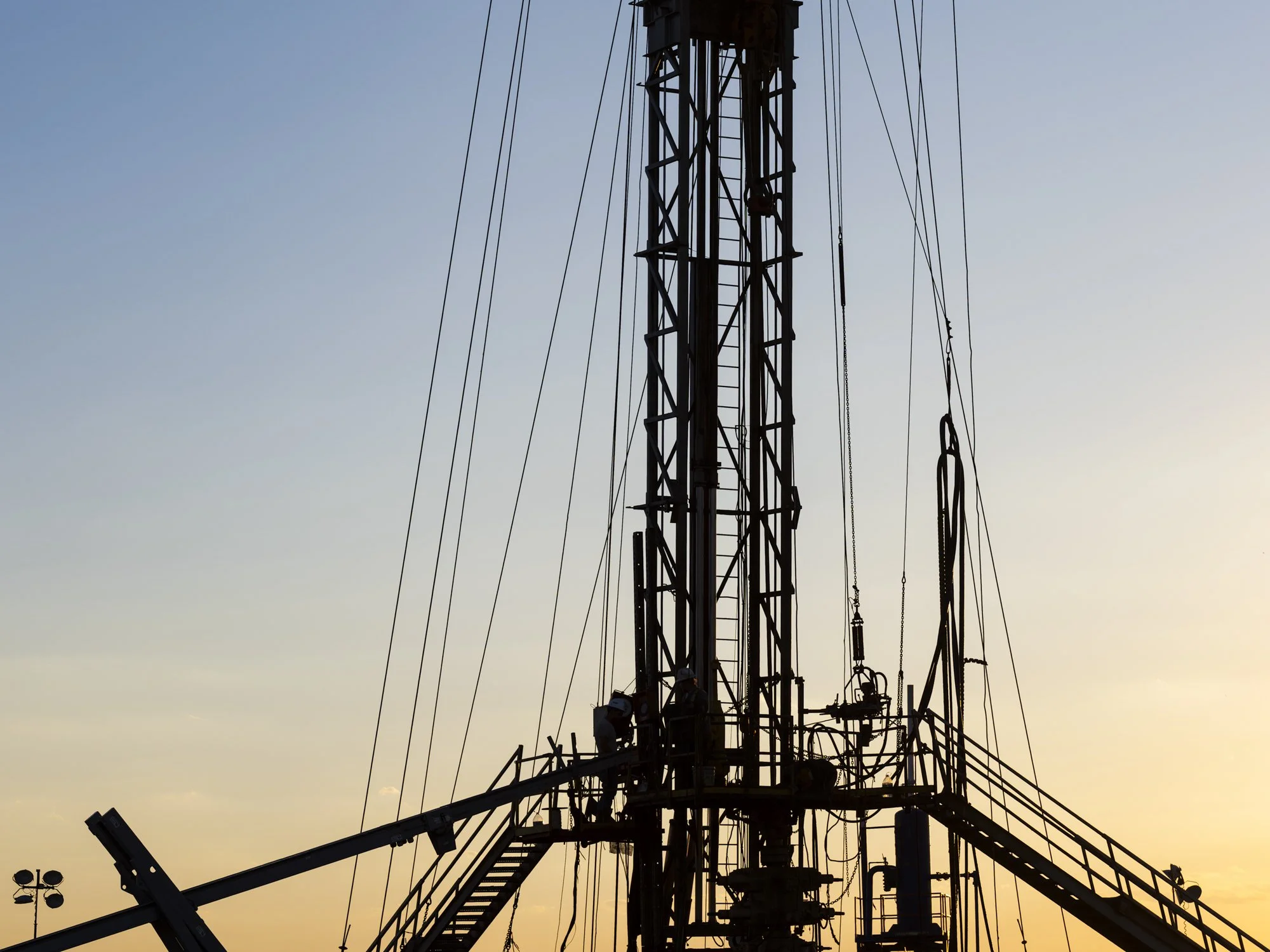 Silhouette of a tall oil or gas drilling rig against a sunset sky with wires and platform details.