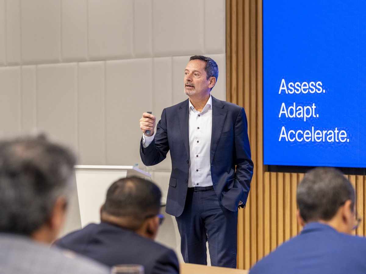 A man in a suit giving a presentation in a conference room, with a screen displaying the words "Assess. Adapt. Accelerate."