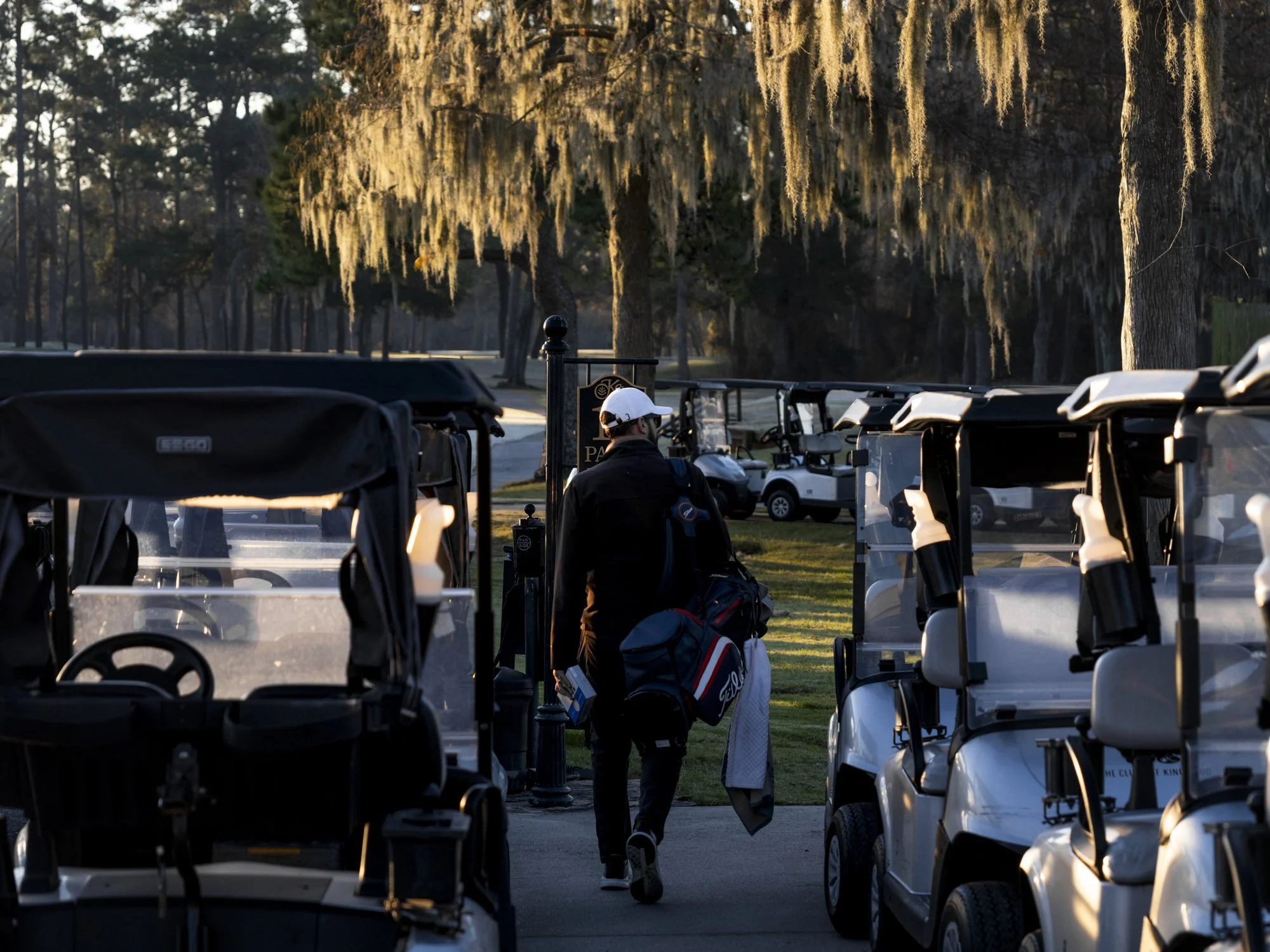 A person with a white cap carrying golf clubs walks between golf carts on a golf course, with trees in the background during sunset.