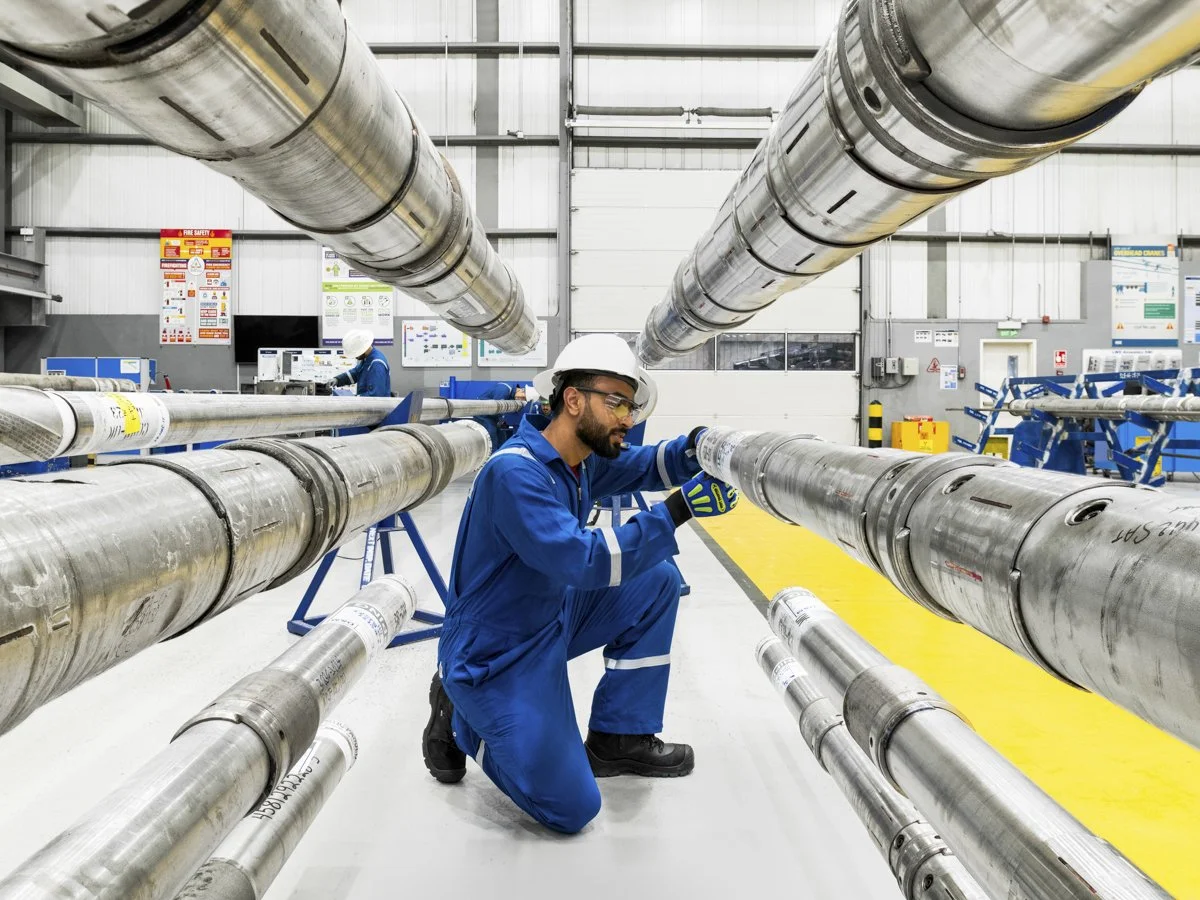 A worker in a blue uniform, safety helmet, and gloves inspects large metal pipes inside an industrial facility, with more pipes and safety signs visible in the background.