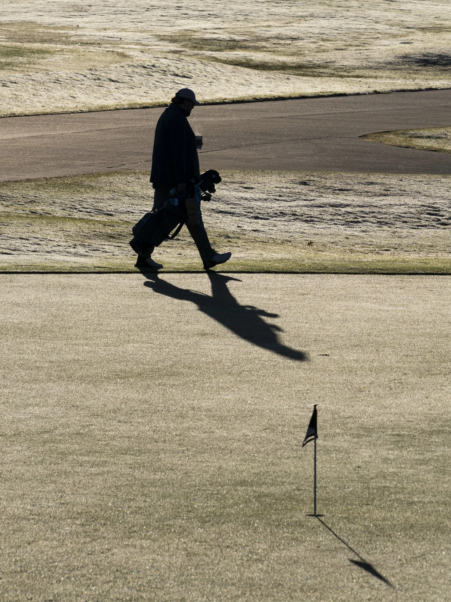 A person walking on a golf course with a golf bag, casting a shadow on the putting green near a hole with a flagstick.