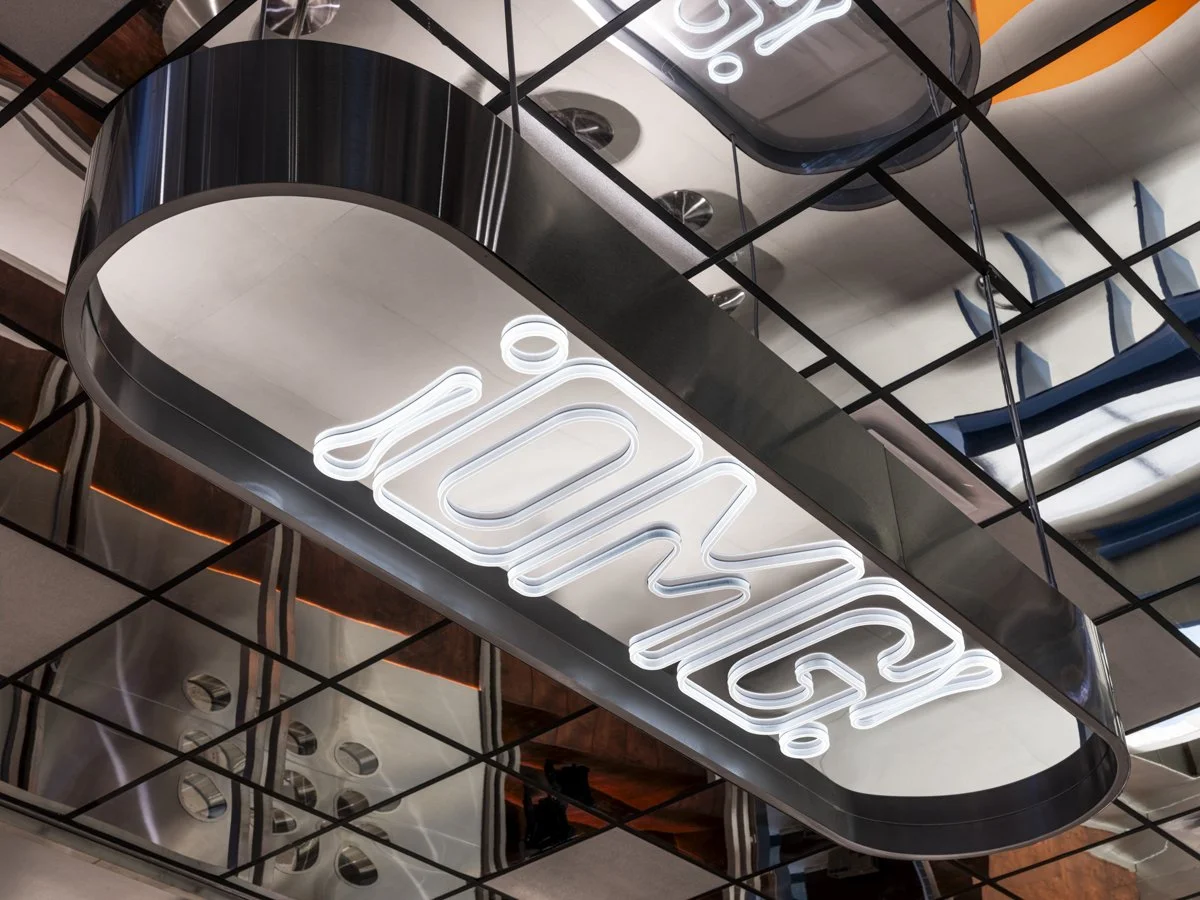 Bright white neon illuminated sign spelling 'NOW' hanging from a grid-like ceiling with reflective black and mirror panels.