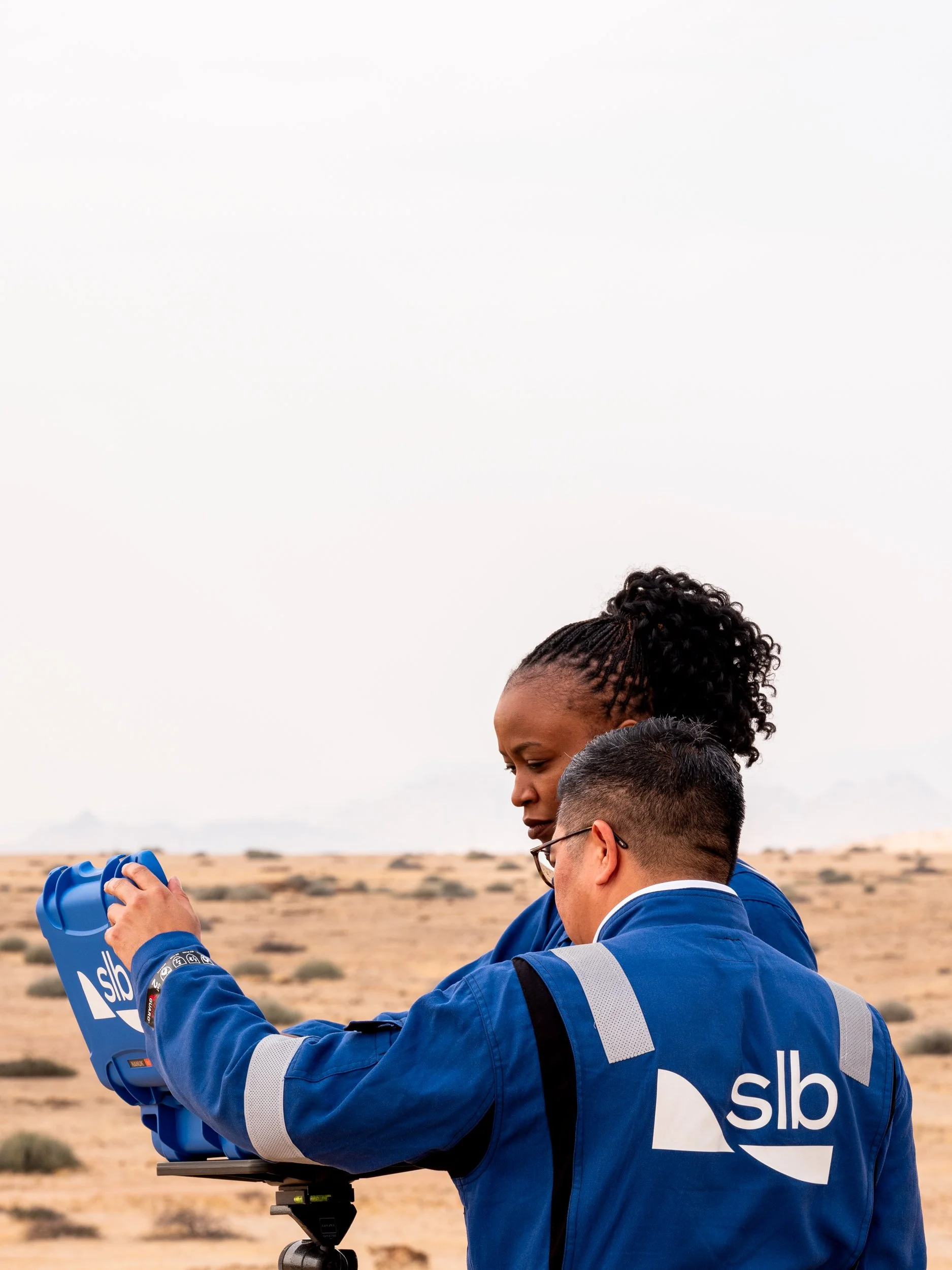 Two scientists in blue lab coats working outdoors in a desert landscape, examining a blue device.