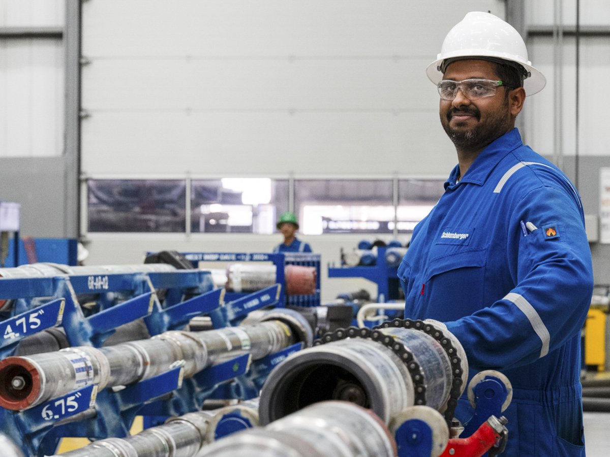 A man in a white safety helmet and clear safety glasses standing in an industrial warehouse, wearing a blue uniform, holding a large industrial roller. In the background, another worker is seen with a green safety helmet, near machinery.