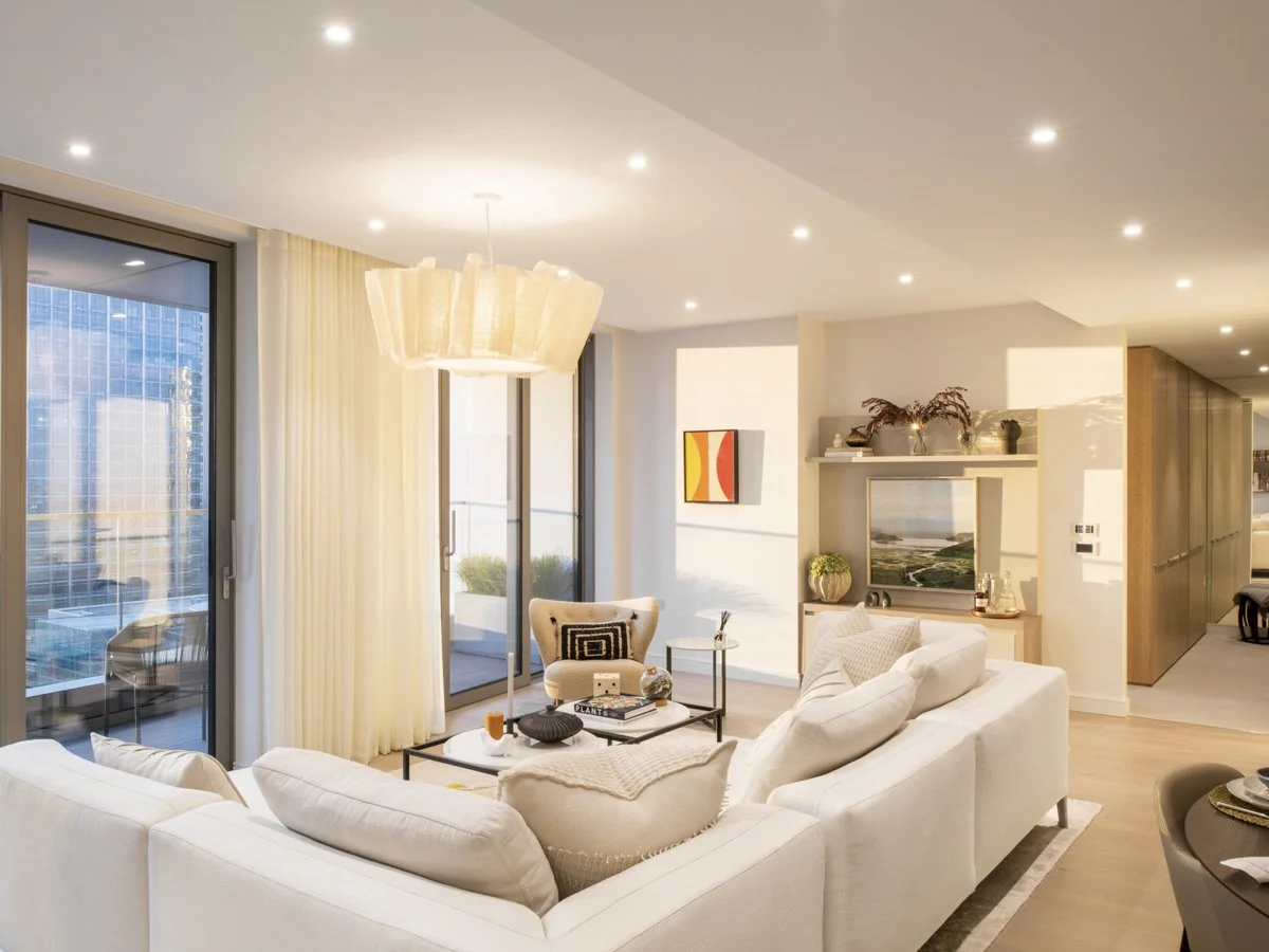 Modern living room with white sofa, armchair, coffee table, large windows with sliding doors, and a flat-screen TV with decorative shelves above.