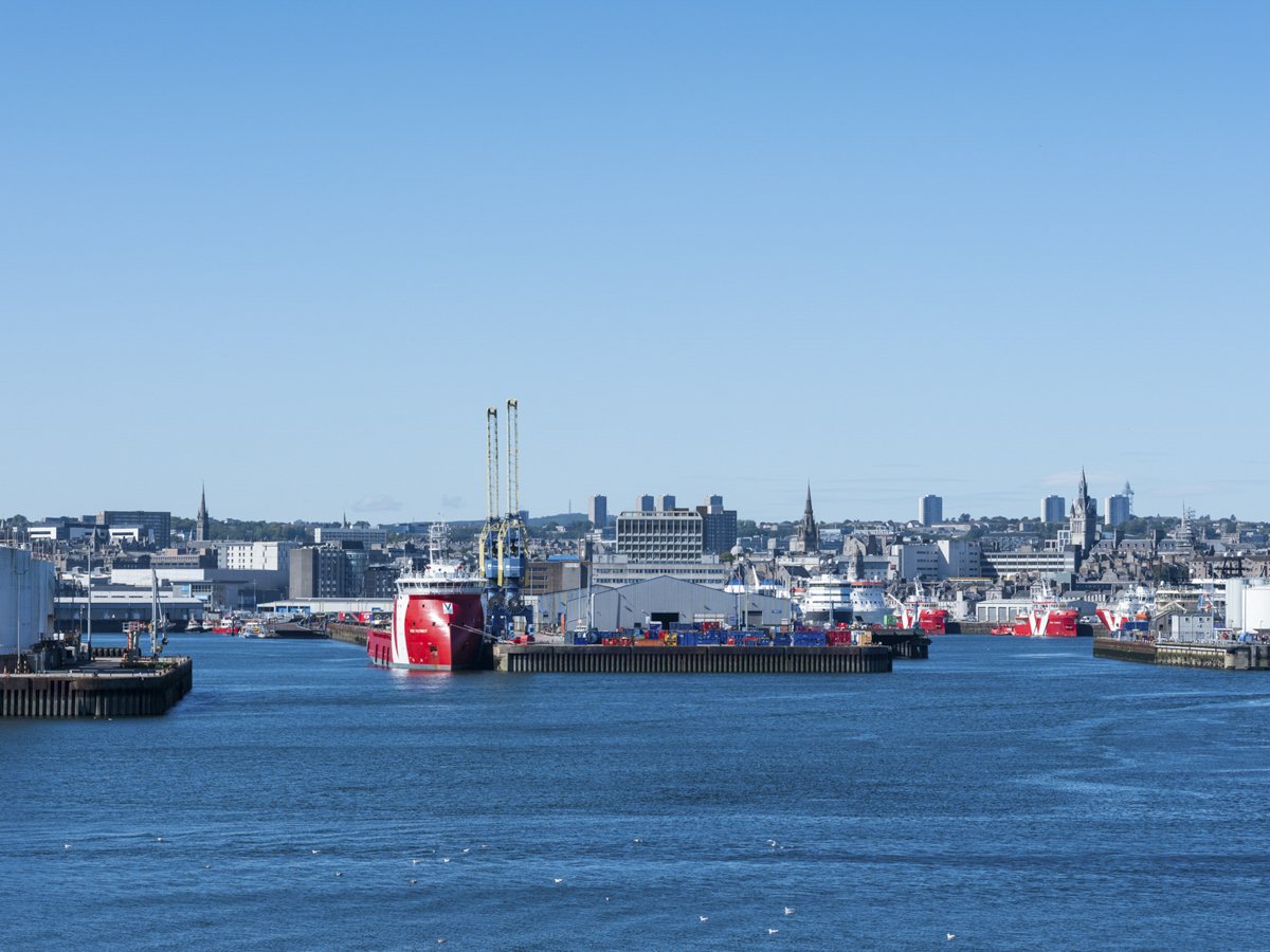 View of a harbor with large ships, cranes, and a city skyline in the background on a clear, sunny day.
