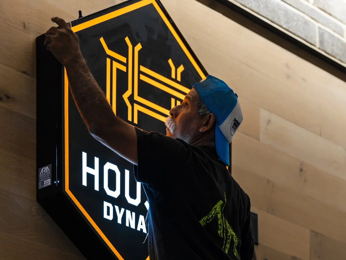 A man installing or adjusting a neon sign that reads 'HOUSE DYNAMIC' on a wooden wall.