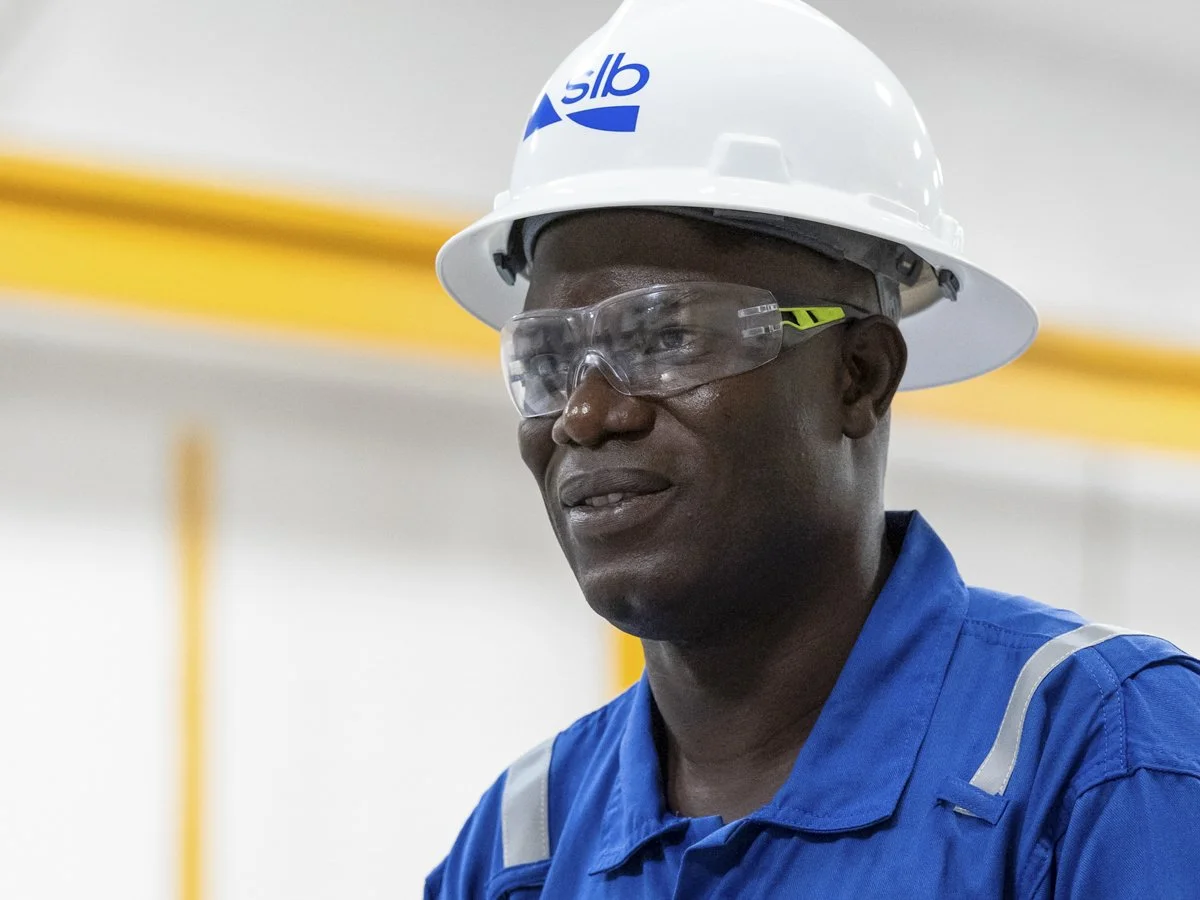 A worker wearing a white hard hat with a logo, safety goggles, and a blue jumpsuit in an industrial setting.