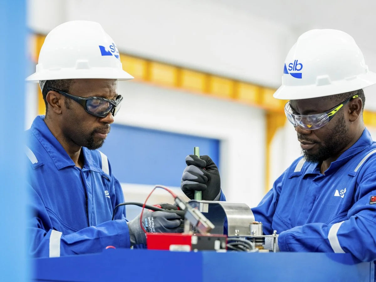 Two male workers in blue uniforms, safety glasses, and white helmets are working on a piece of machinery in an industrial setting.