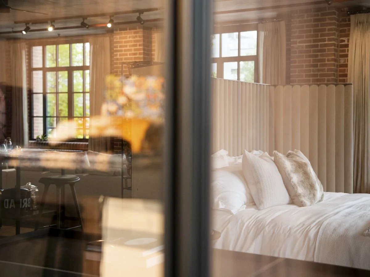 View of a modern bedroom with a large bed, beige headboard, white bedding, and throw pillows, with windows and brick walls in the background.