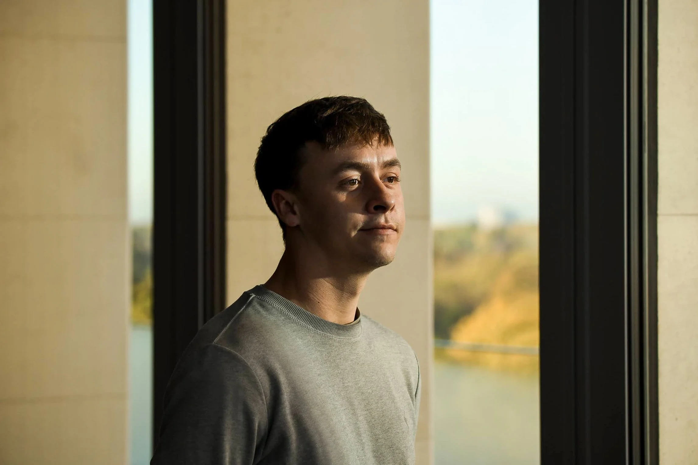 A young man looking out of a window during daytime, with a calm expression and a landscape view in the background.