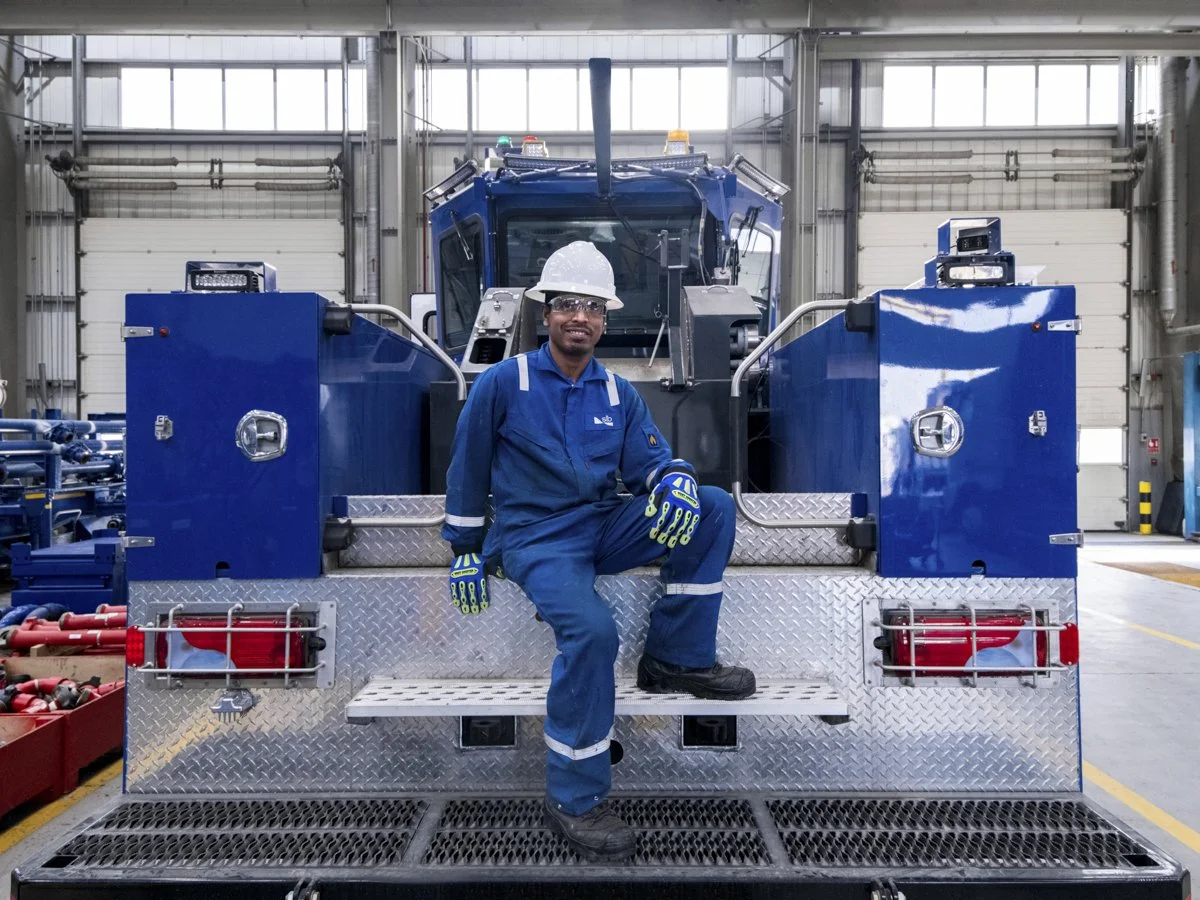 A worker in blue coveralls, safety gloves, helmet, and glasses sitting on a step in front of a large blue industrial vehicle inside a warehouse or factory.