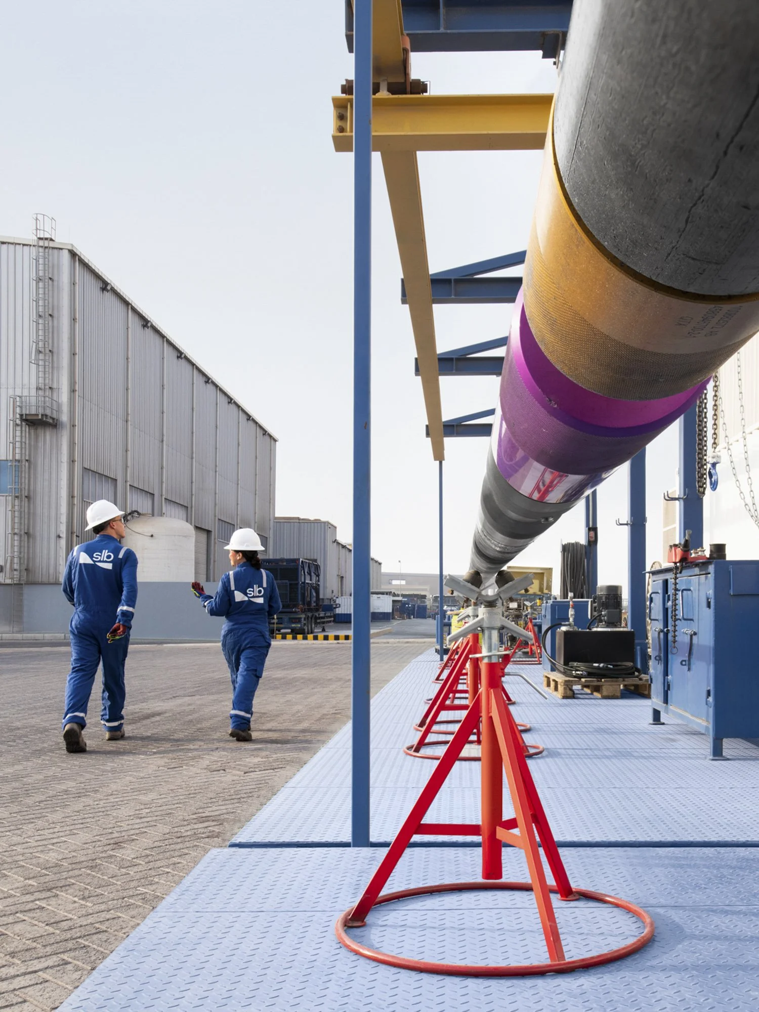 Two workers in blue uniforms and white helmets walking beside a large pipeline at an industrial site.