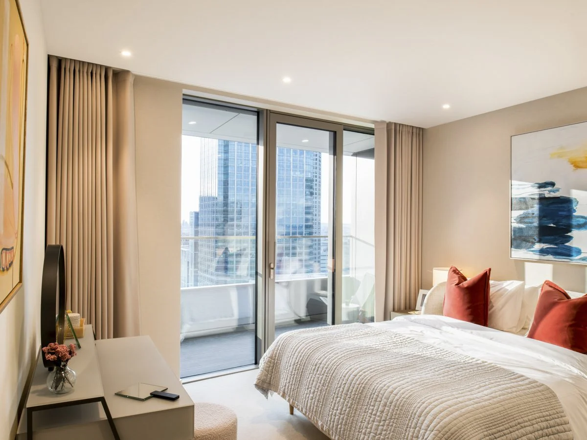 Modern bedroom with large sliding glass door leading to balcony, city view with tall buildings, beige curtains, bed with white bedding and red pillows, wall art, and a small desk with a TV and decorative items.