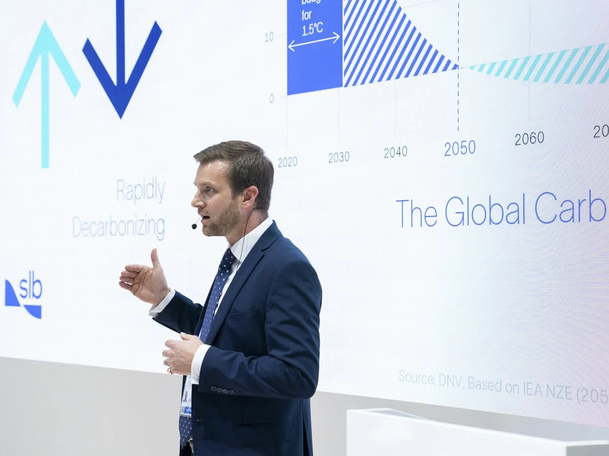 Man in a suit giving a presentation with a large screen displaying a graph titled 'The Global Carbon' and phrases 'Rapidly Decarbonizing'.