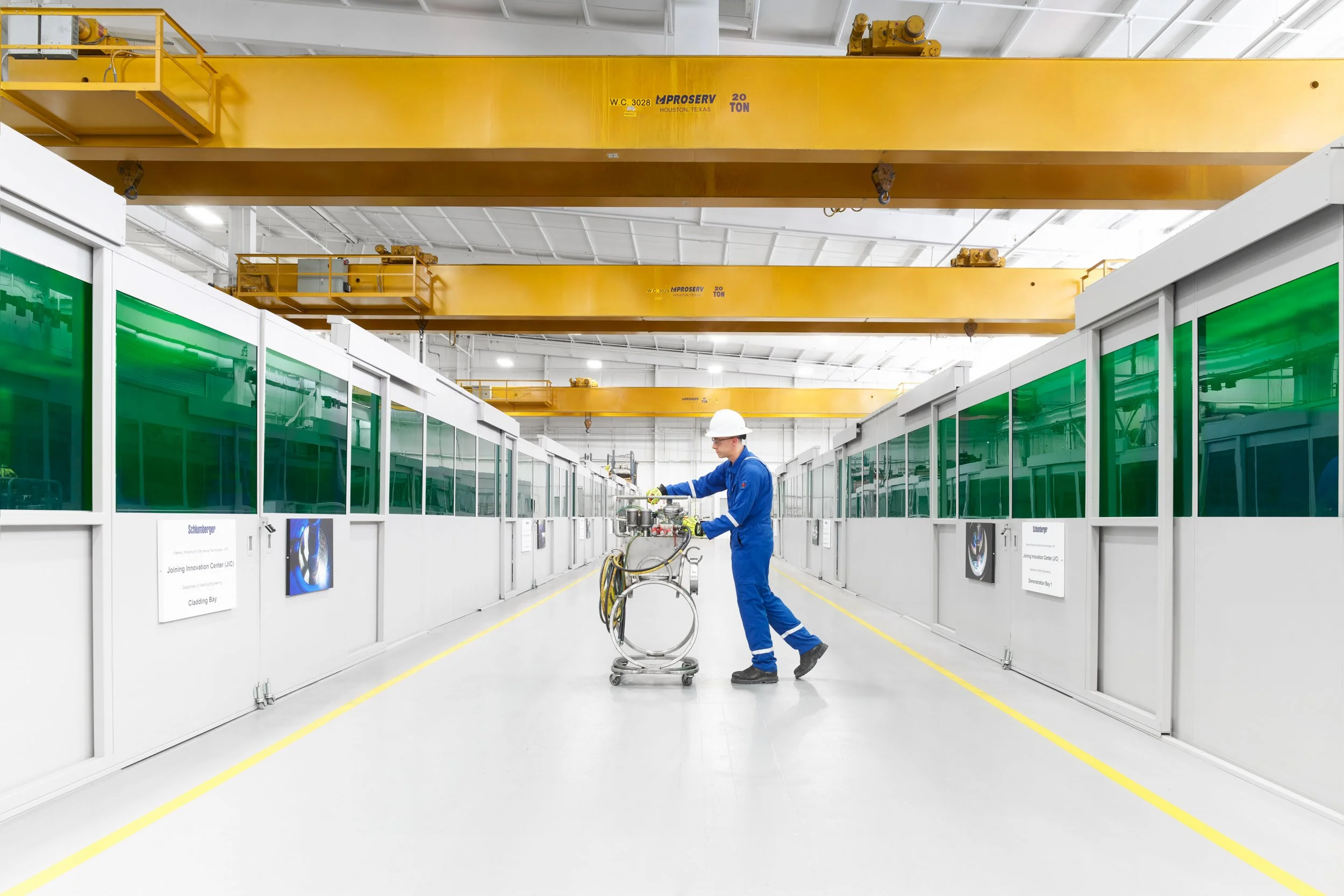 A worker in blue coveralls and a white safety helmet operating machinery in a clean, industrial manufacturing facility with yellow overhead cranes.