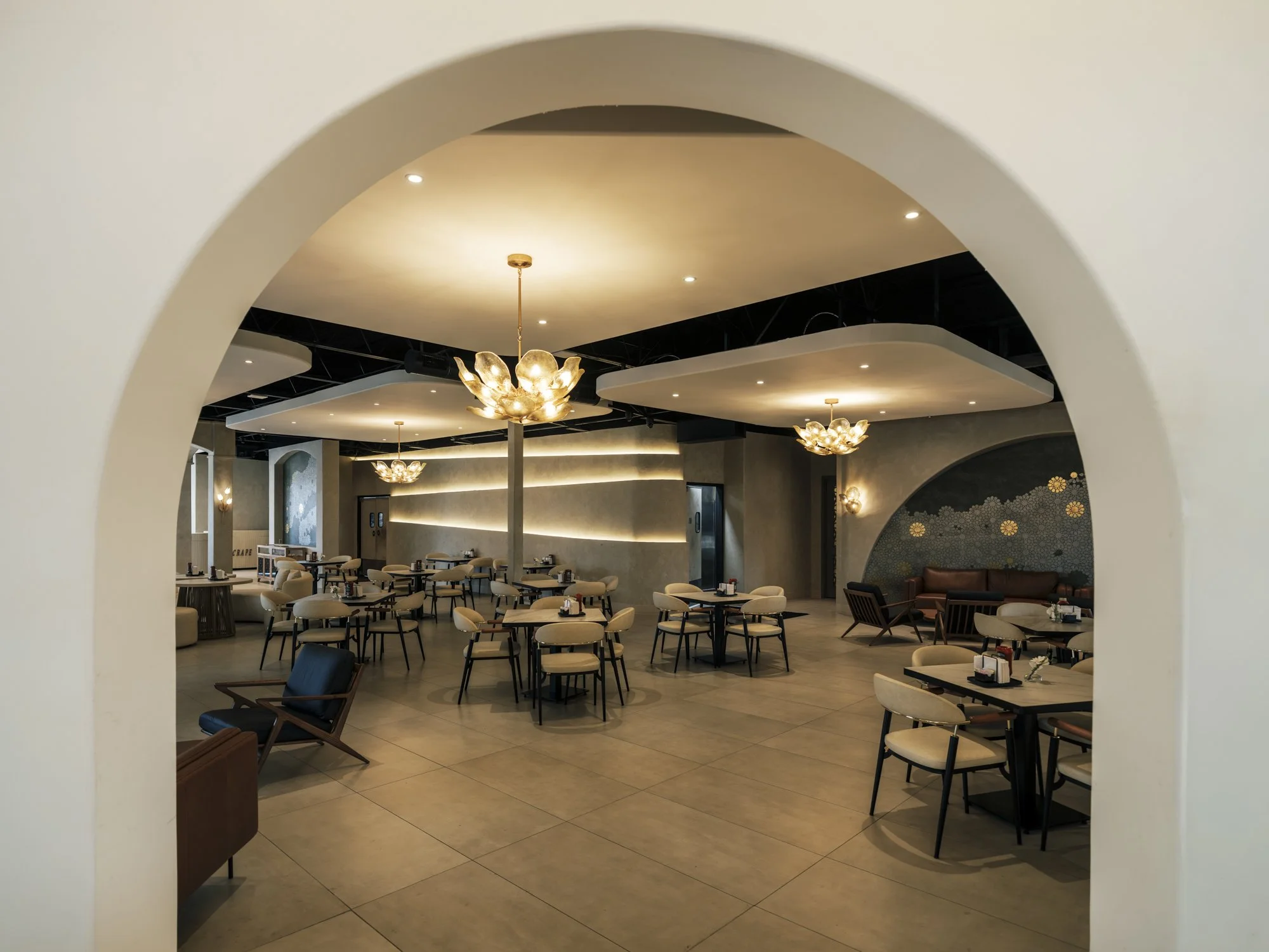 Elegant restaurant interior with modern chandeliers, beige chairs, and dark tables, viewed through an arched doorway.