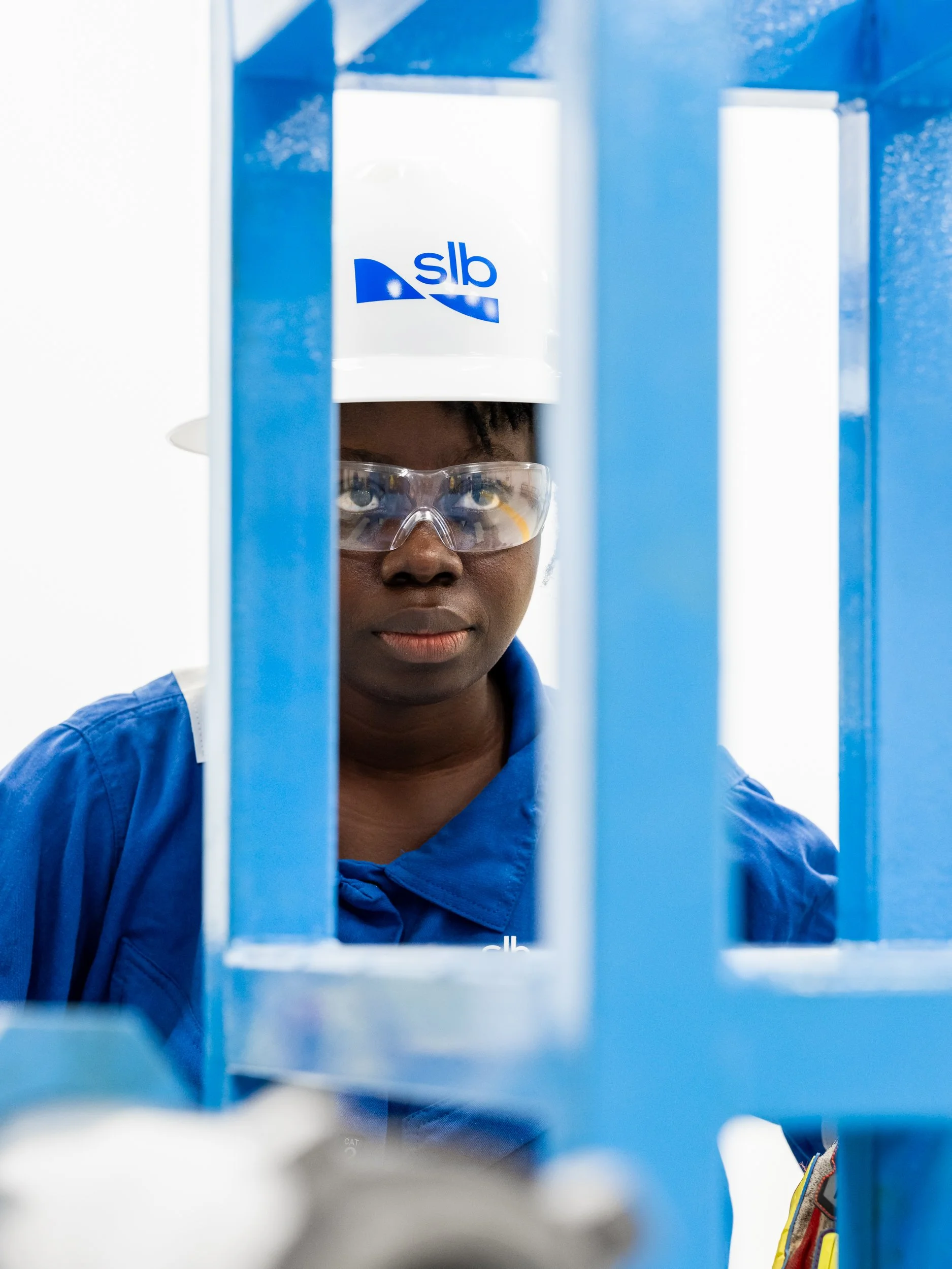 A female scientist wearing safety glasses, a white hard hat with blue logo, and a blue shirt, looks through blue metal bars in a laboratory setting.