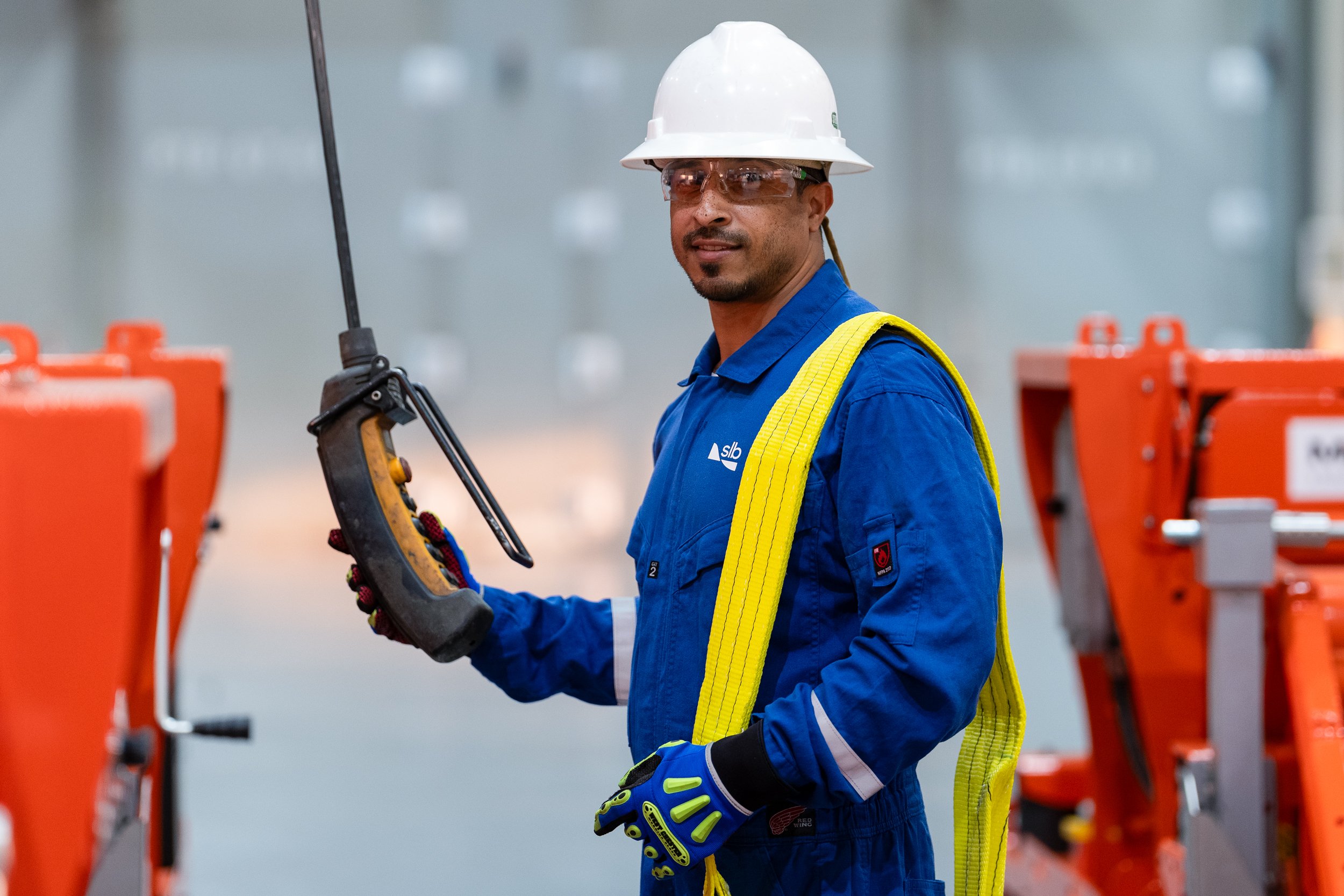 A male worker in safety gear holding a long tool in an industrial setting with orange equipment around him.
