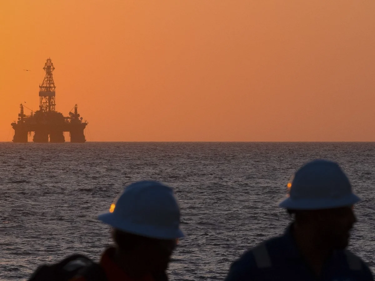 Silhouettes of two workers wearing hard hats on a ship, with an offshore oil rig in the distance at sunset.