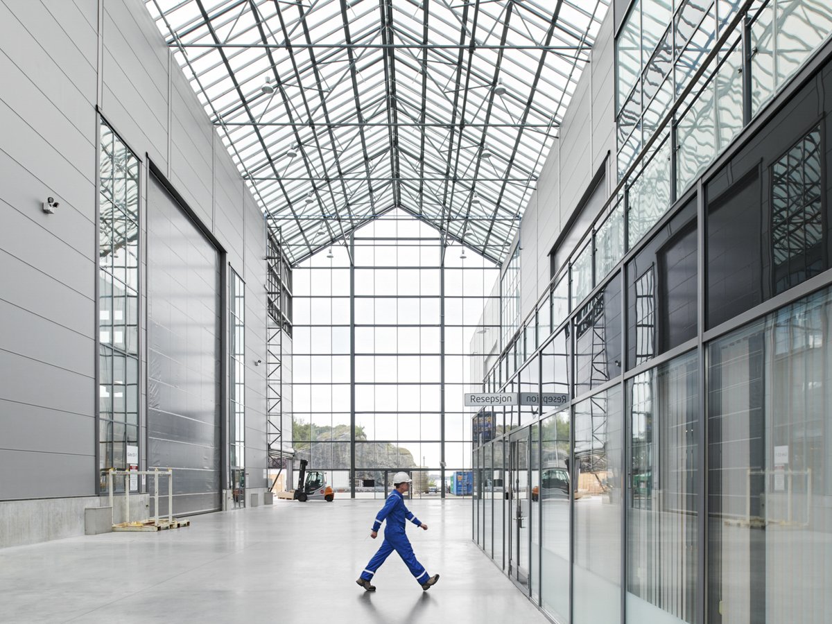 Interior of a modern building with a high glass ceiling, large glass windows, and a person in blue work clothes and a white helmet walking inside.