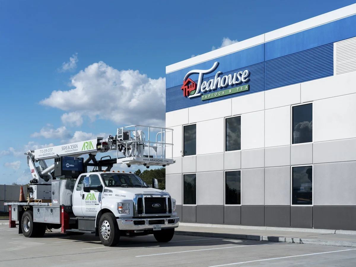 A truck with a cherry picker lift parked outside a building with a sign that reads 'Teahouse Tapioca & Tea' against a blue sky with clouds.