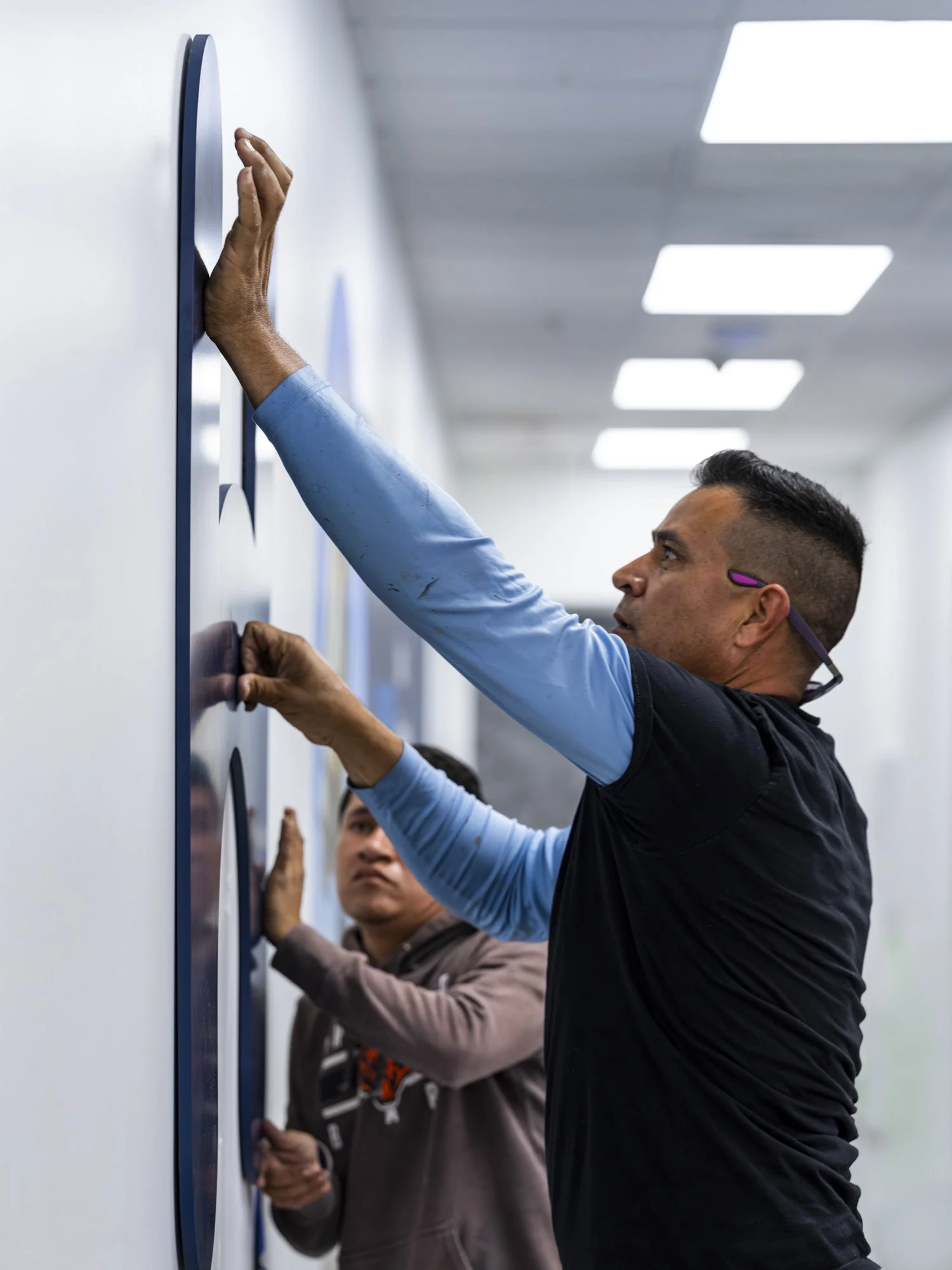 Two men installing or adjusting a large display or wall art on a white wall in a brightly lit corridor.